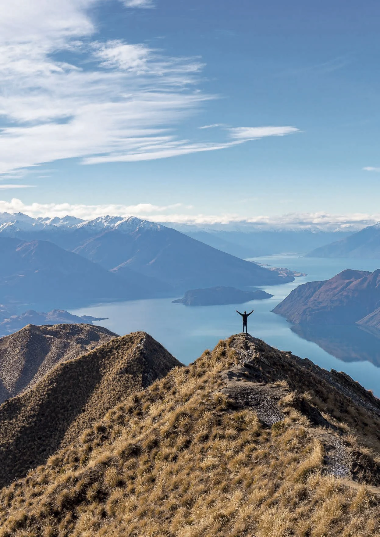 Person standing with arms raised on a mountain ridge overlooking a lake and mountain range under cloudy sky.