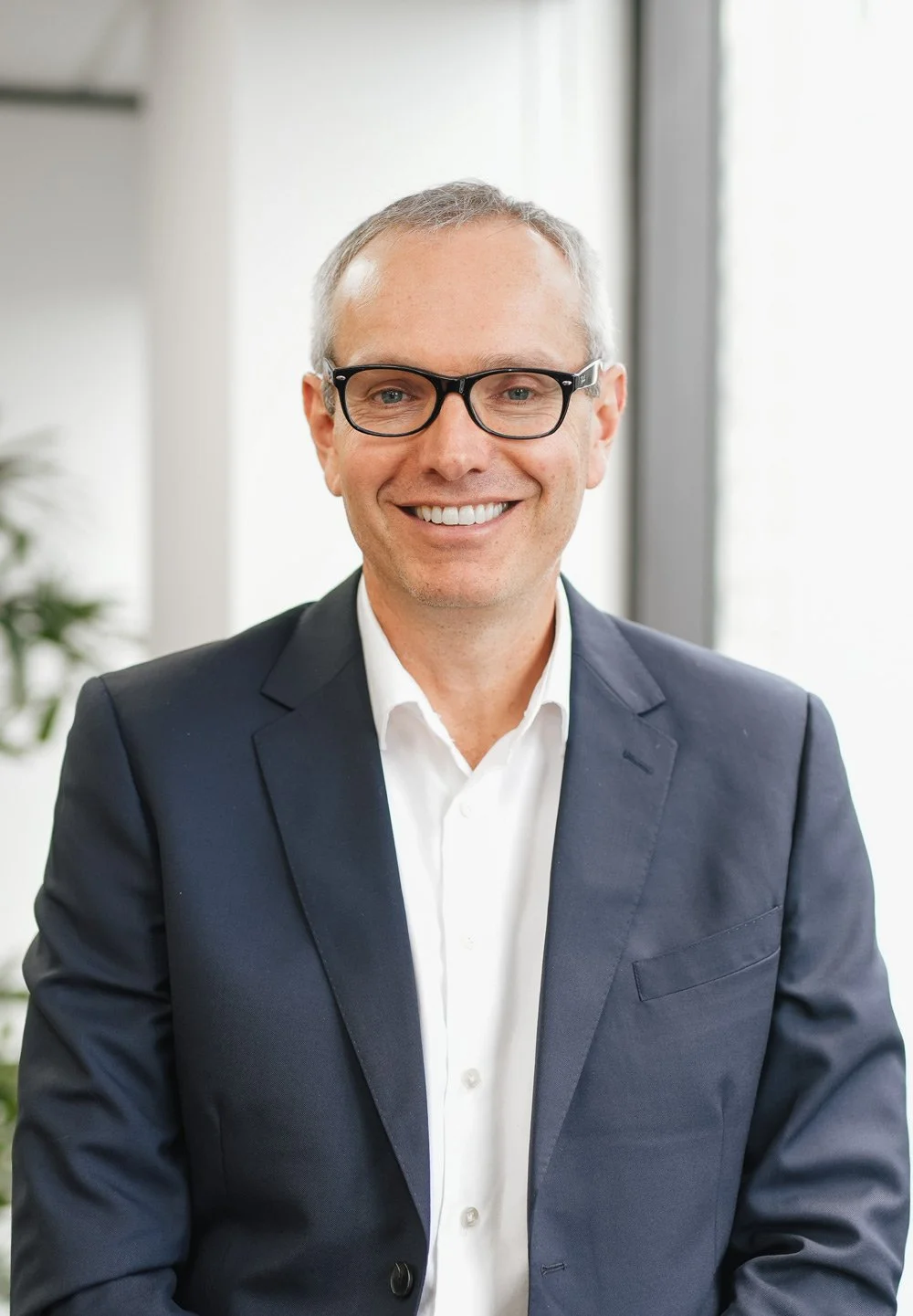 Portrait of a smiling middle-aged man with glasses, wearing a dark suit and white shirt, in a bright office setting with large windows in the background.
