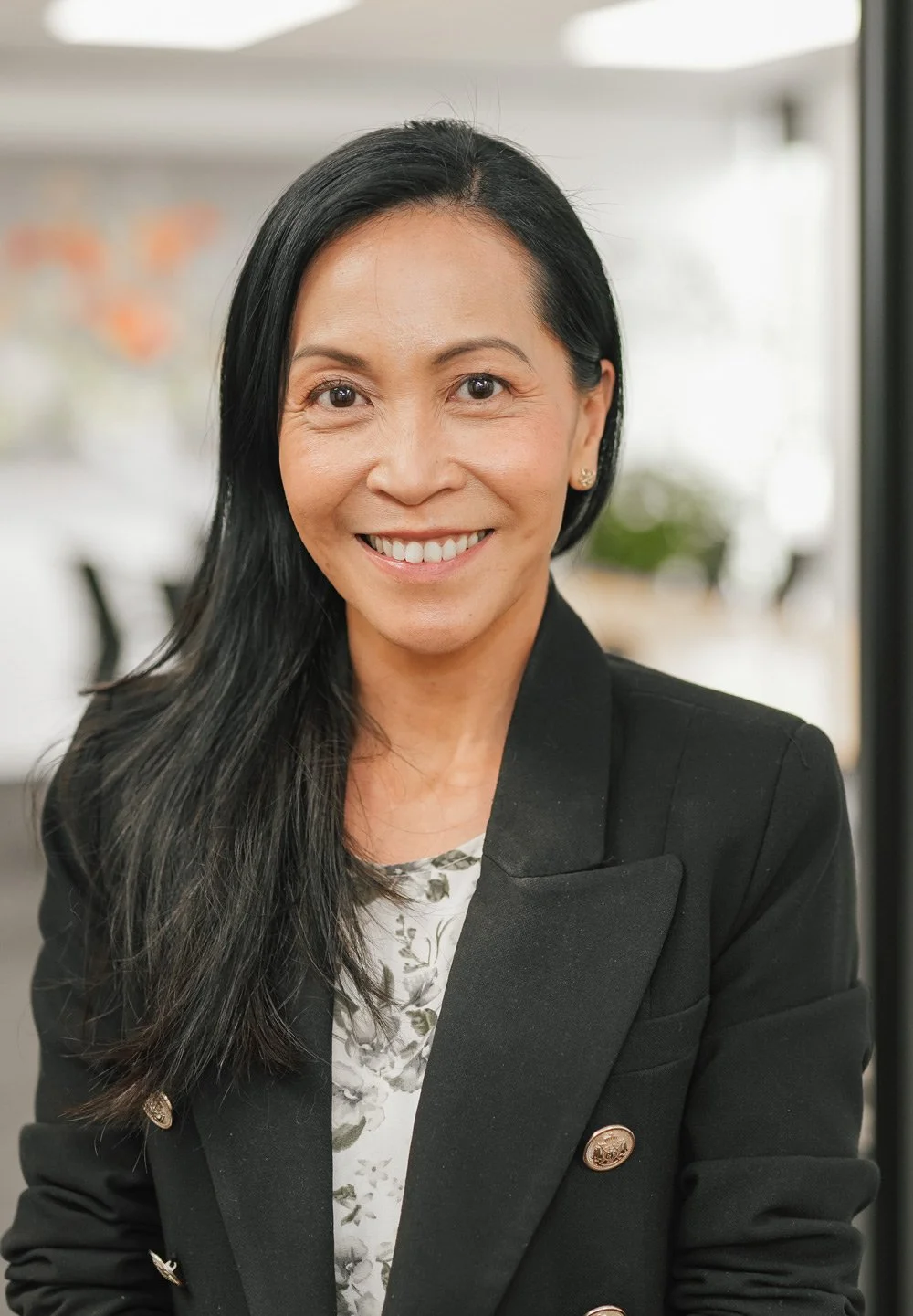 A professional woman with long black hair smiling, wearing a black blazer with gold buttons and a floral top, in an indoor setting with blurred background.