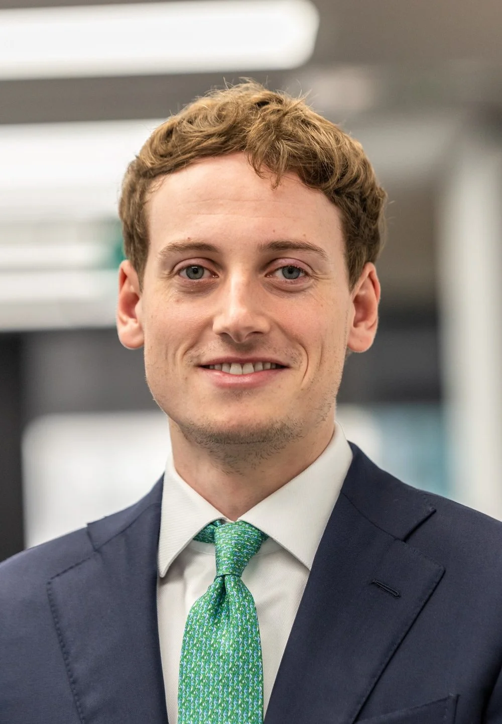 A young man with light skin, short red hair, and blue eyes, wearing a dark suit, white shirt, and a green patterned tie, smiling in a professional setting.