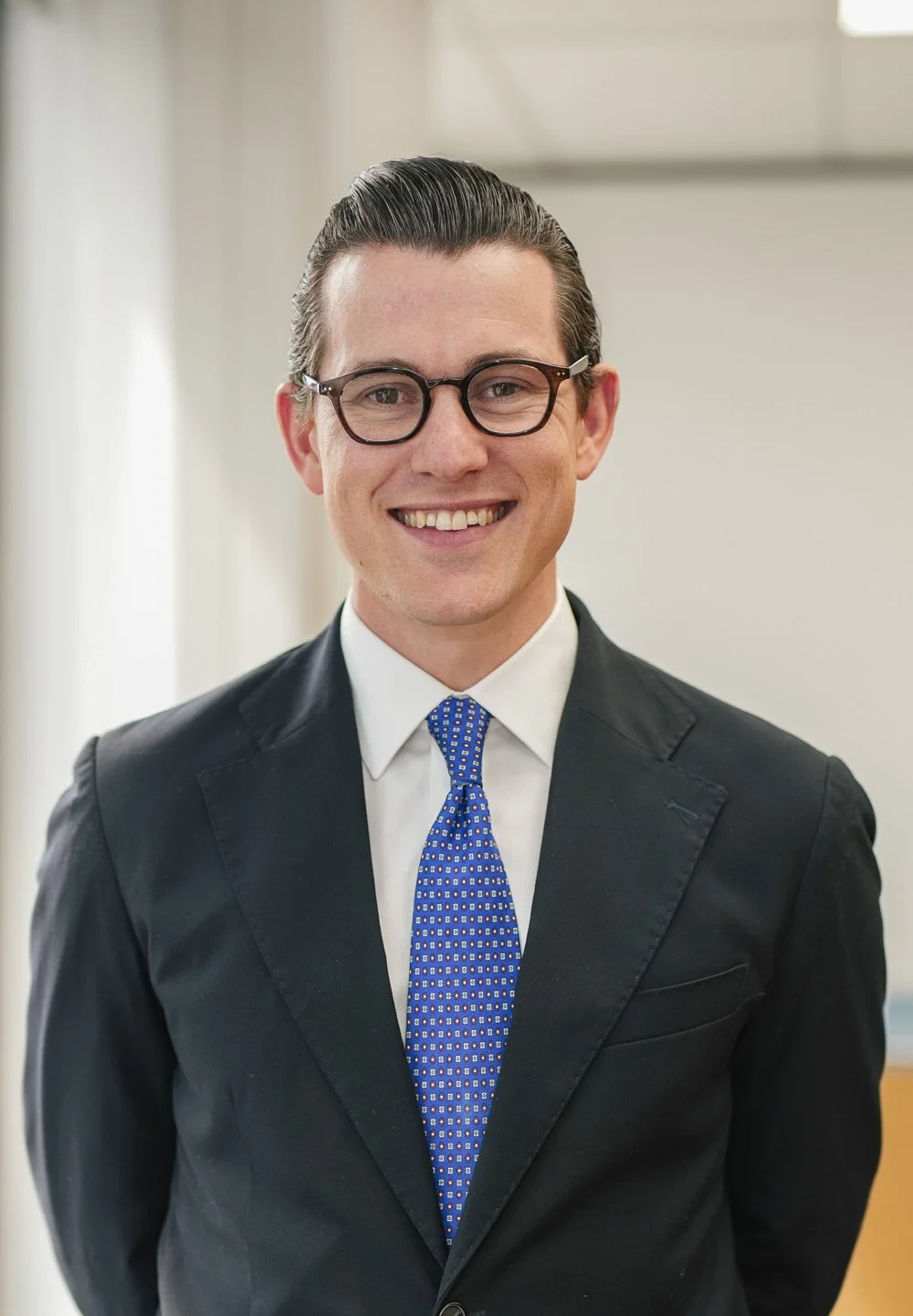 A smiling man in a business suit with glasses, white shirt, and blue patterned tie, standing indoors with a neutral background.