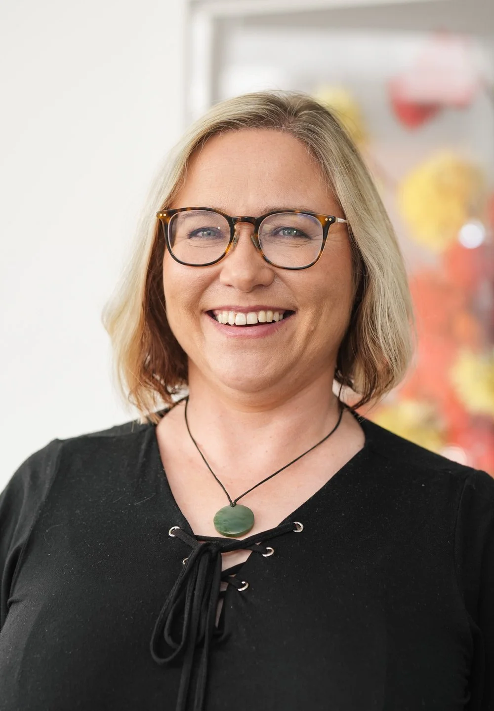 Smiling woman with blonde hair, glasses, and a black top, standing indoors with a colorful blurred background.