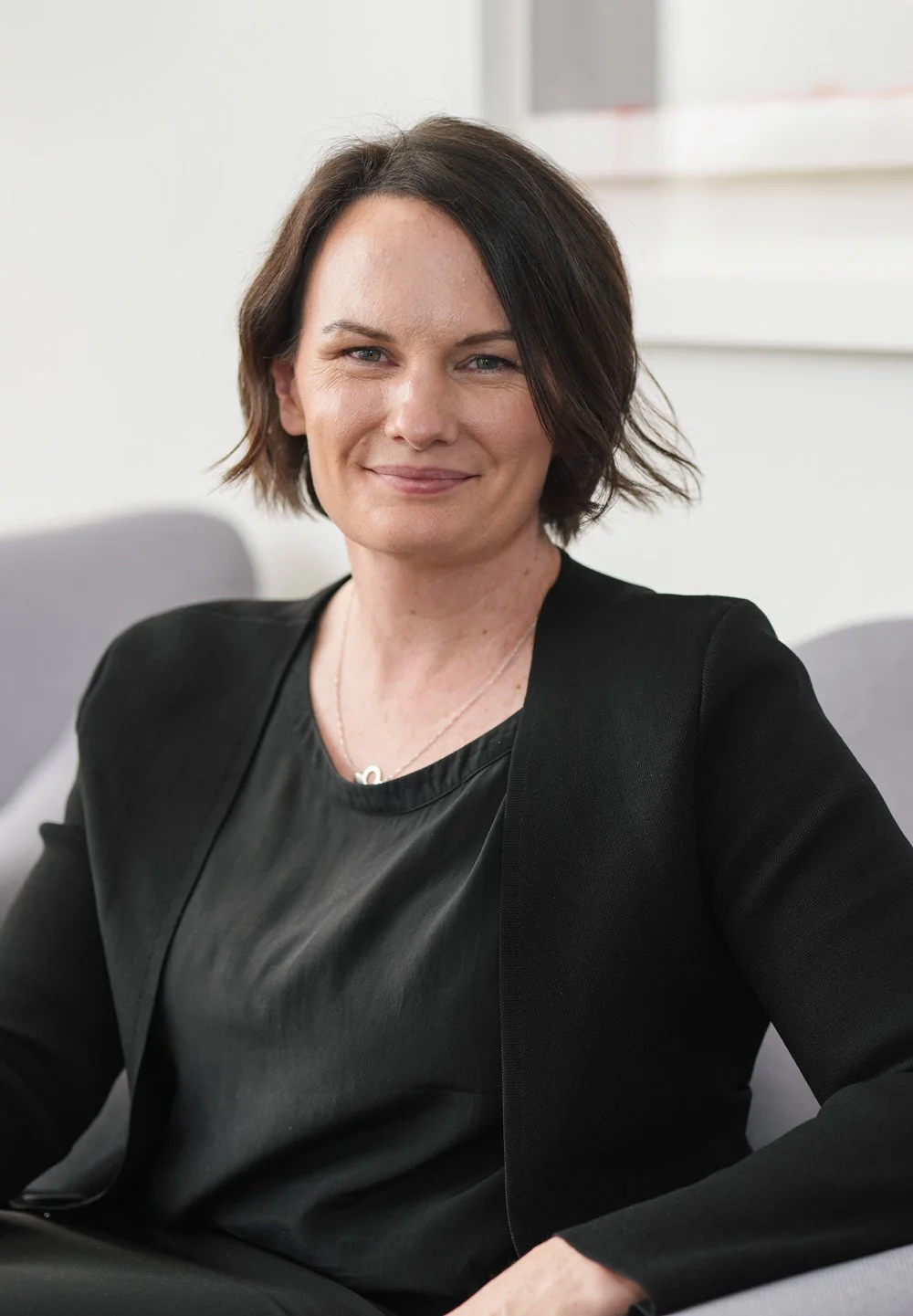 A woman with short dark brown hair styled in a bob, wearing a black blazer and a black top, sitting on a grey chair in a modern office setting, smiling at the camera.