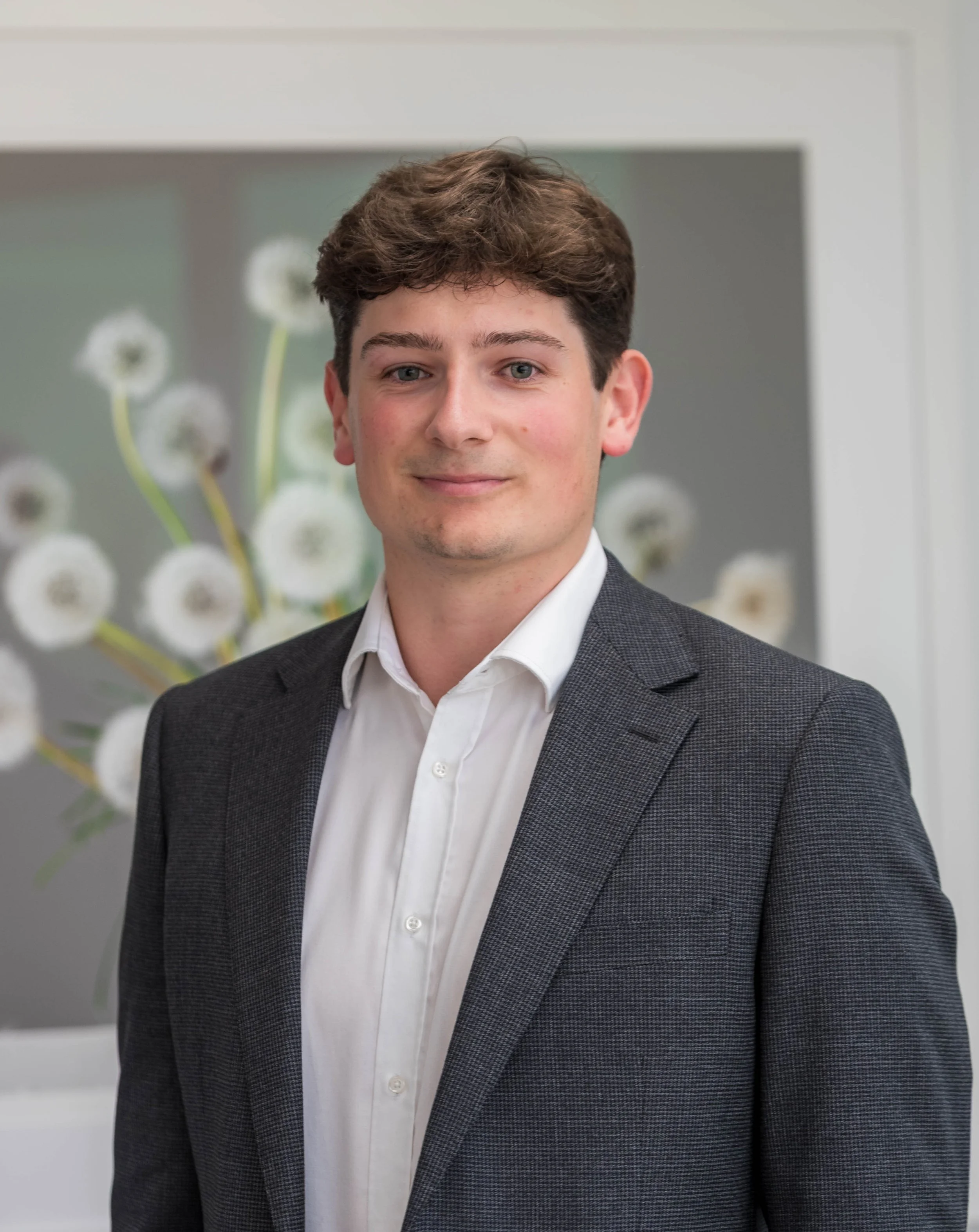 Young man with brown hair, wearing a white shirt and a dark checkered blazer, standing indoors with blurred floral artwork in the background.