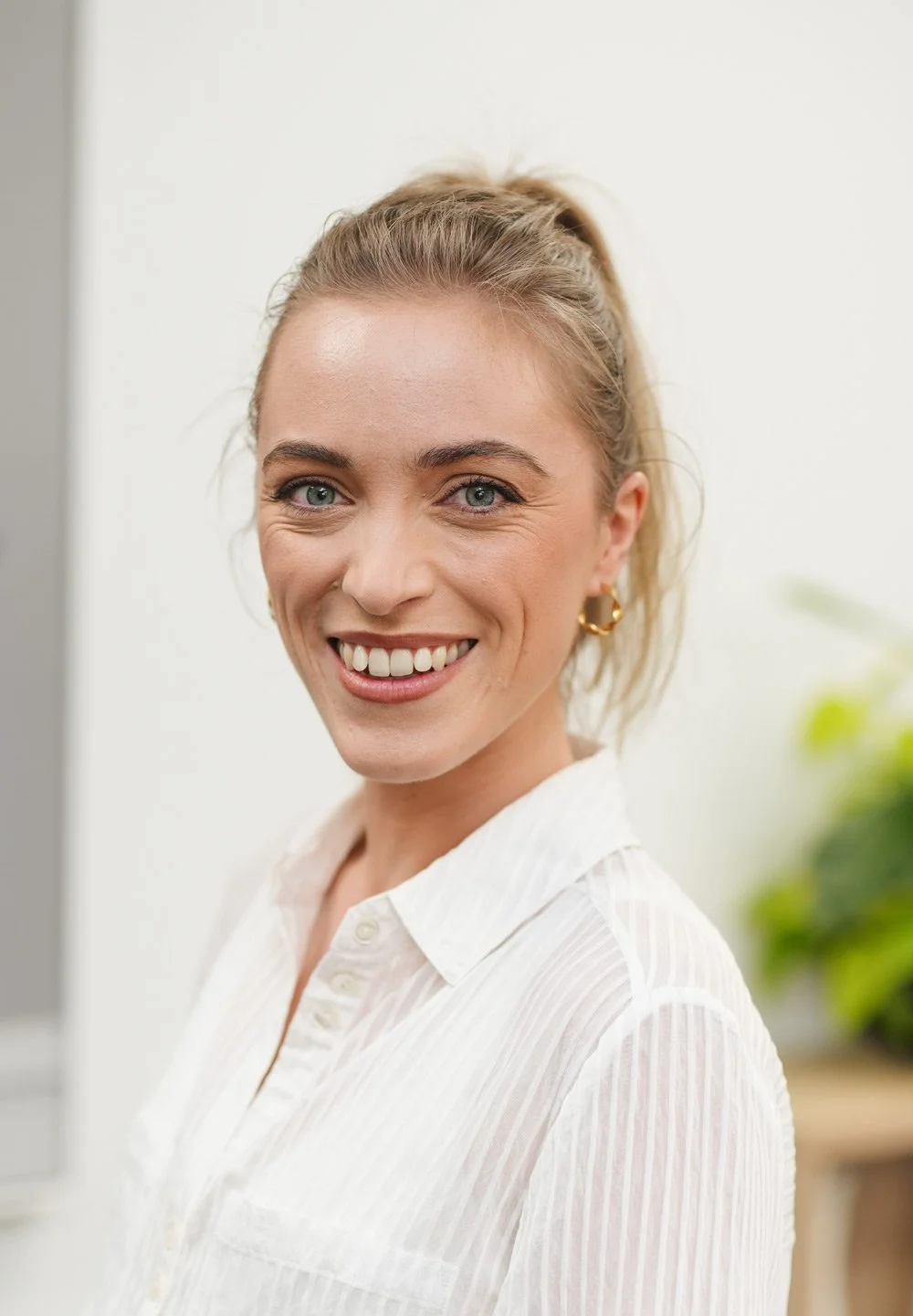 A smiling woman with blonde hair tied in a ponytail, wearing a white button-up shirt and gold earrings, standing indoors with a blurred background of a plant.
