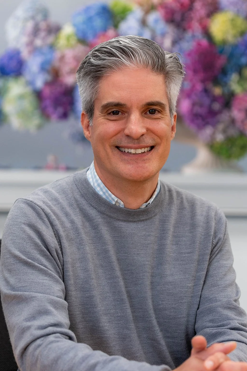 Smiling middle-aged man with gray hair, wearing a gray sweater and a checked shirt, sitting in front of a colorful blurred floral background.