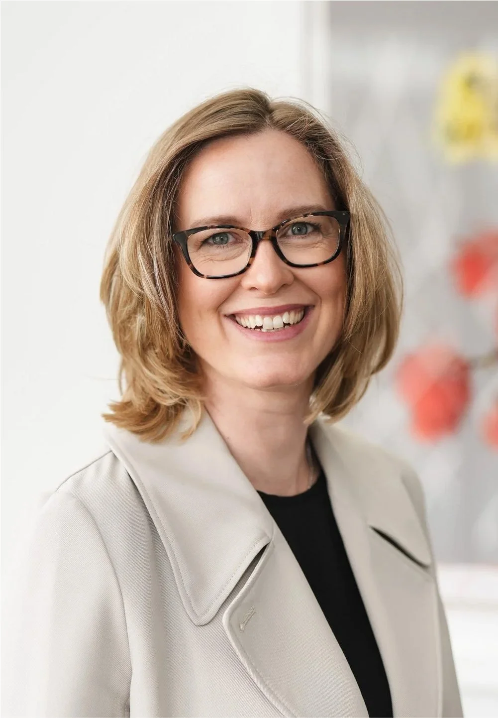 A woman with shoulder-length blonde hair, glasses, and a bright smile, dressed in a light-colored blazer and a black top, standing indoors with a blurred background of colorful objects.