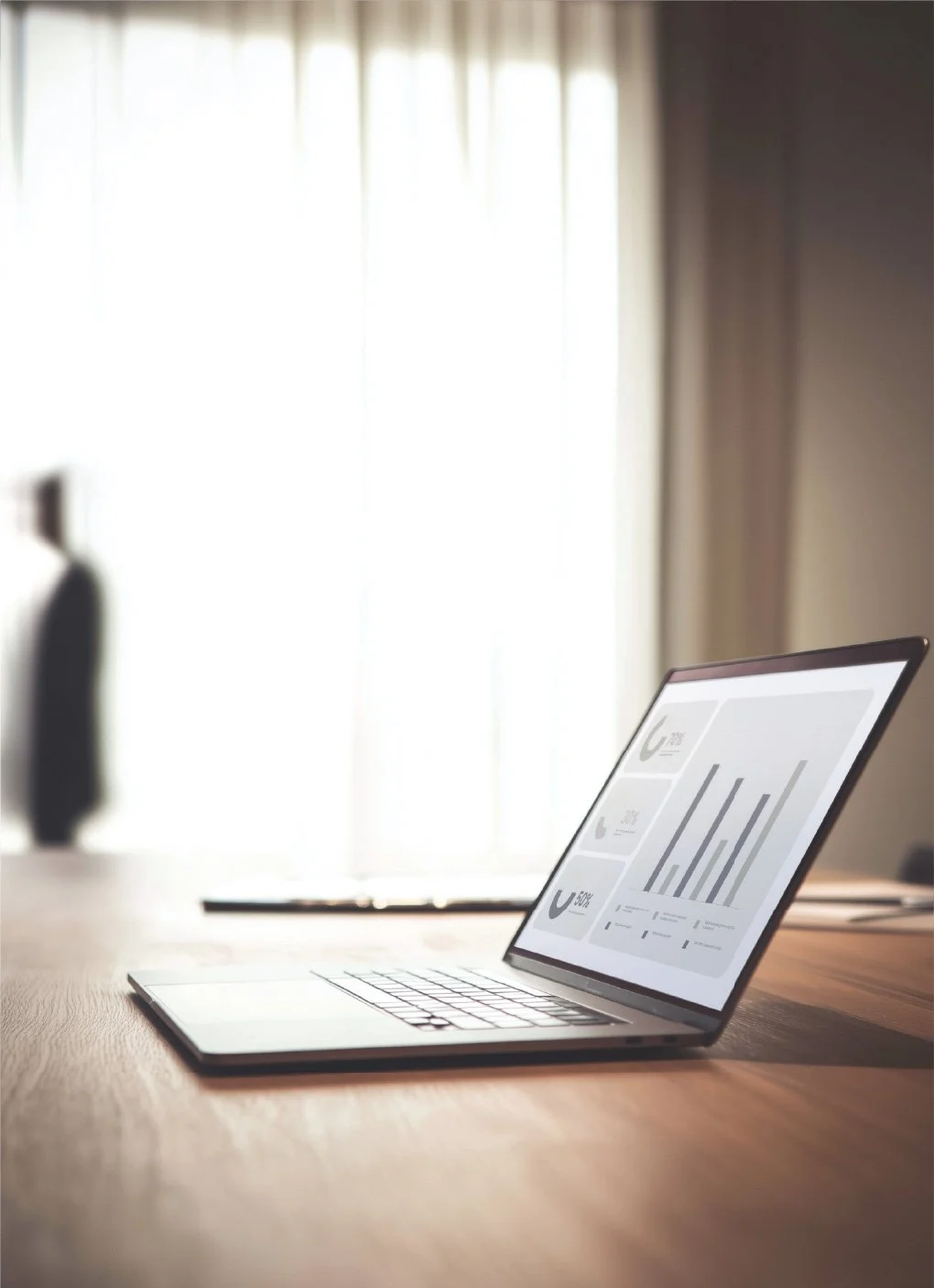 A laptop on a wooden desk with a bright window in the background, displaying graphs and data on its screen.