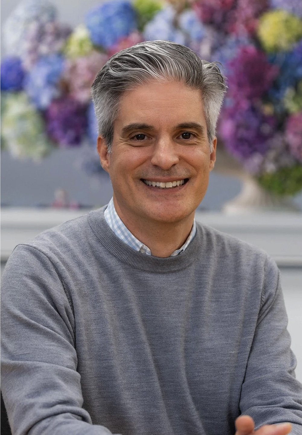 A smiling middle-aged man with salt-and-pepper hair, wearing a gray sweater over a checkered shirt, sitting in front of a colorful blurred floral background.
