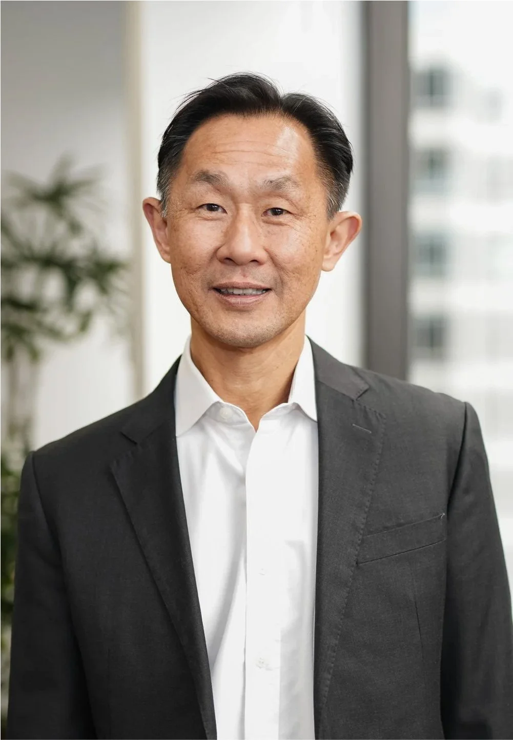 Professional Asian man in a dark suit and white shirt standing indoors in front of a window with a city view.