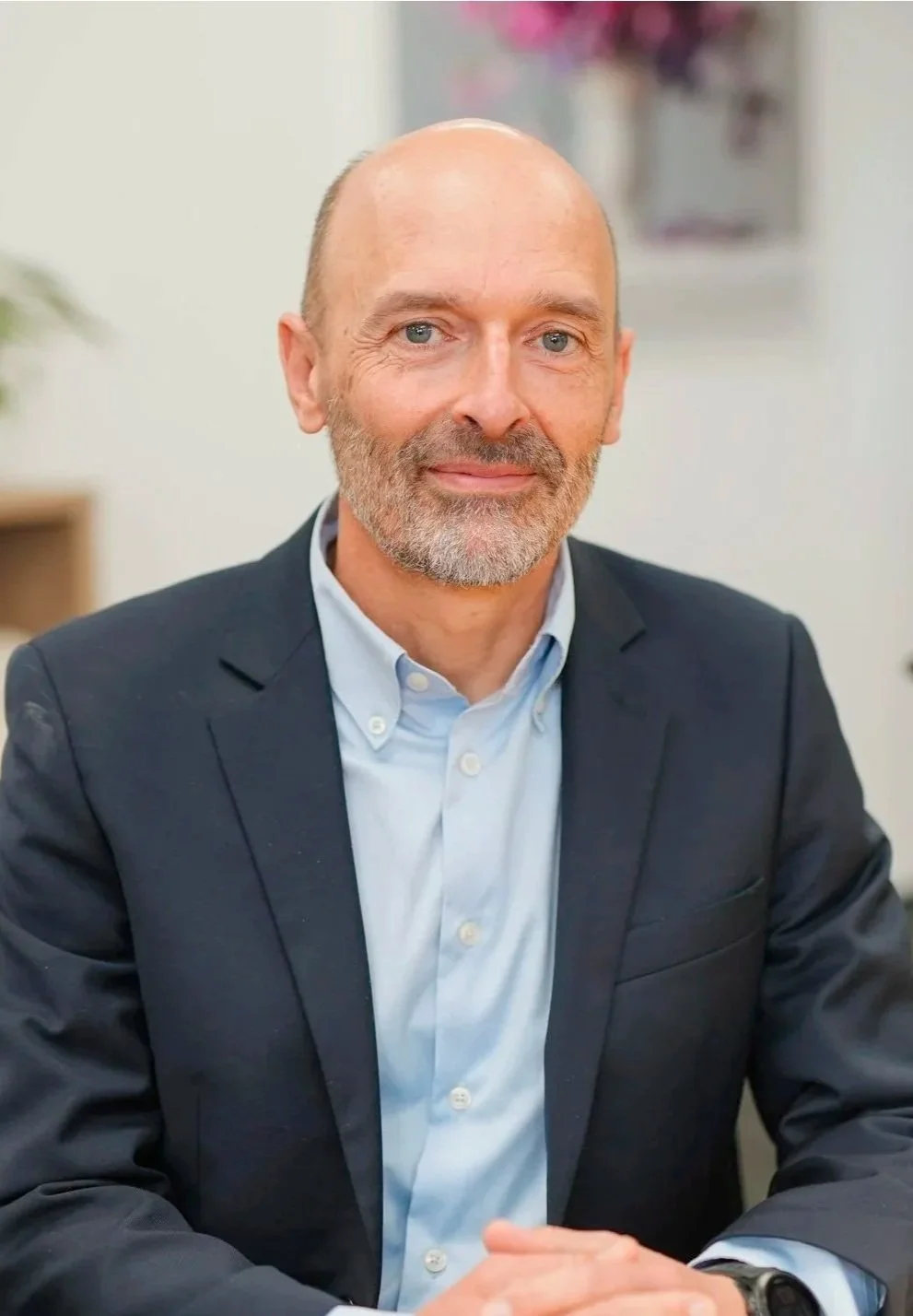 A middle-aged man with a beard and balding head, wearing a dark suit jacket and a light blue dress shirt, sitting at a table in an office setting.