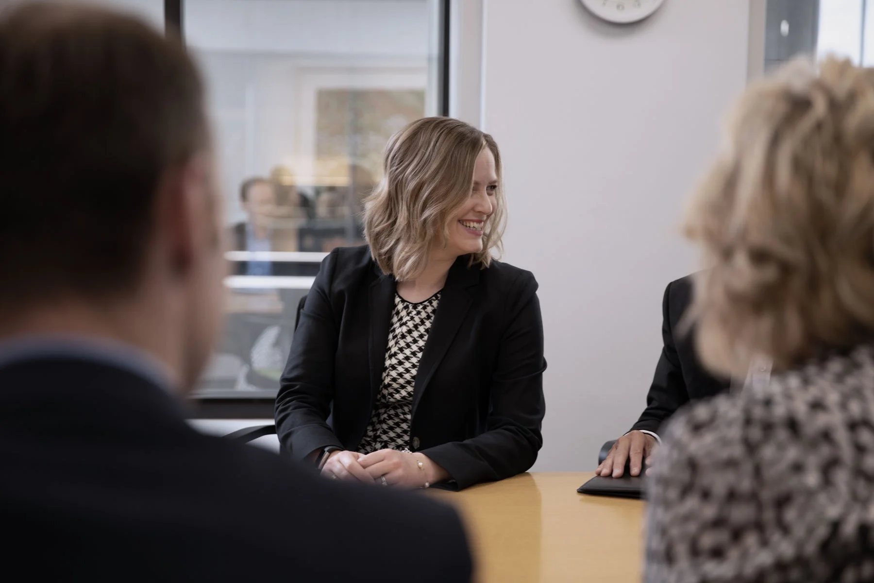 A woman with shoulder-length wavy blond hair smiling during a business meeting with three other people, all sitting around a conference table in a modern office.