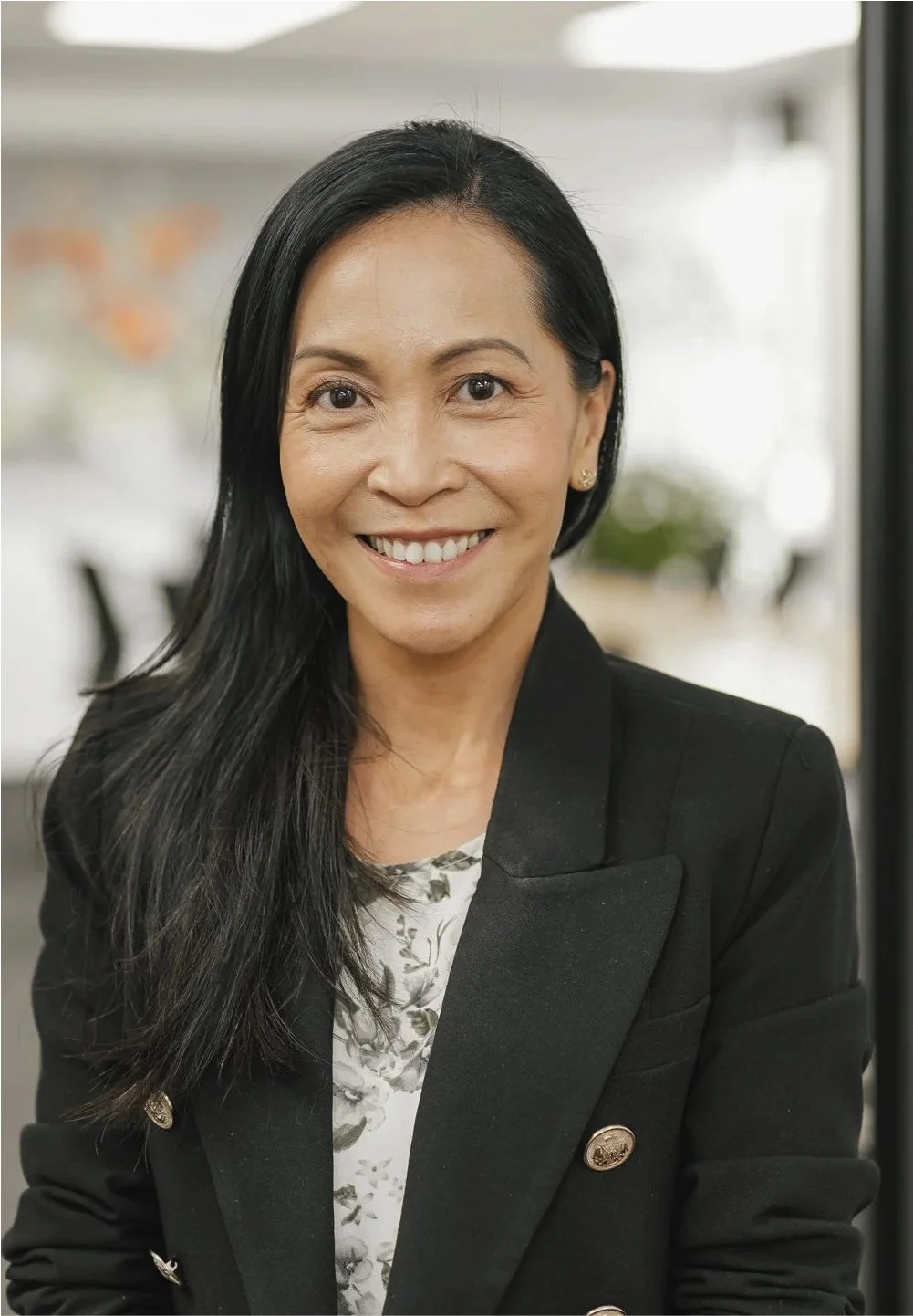 A smiling woman with long black hair, wearing a black blazer with gold buttons over a patterned blouse, standing indoors near a window.