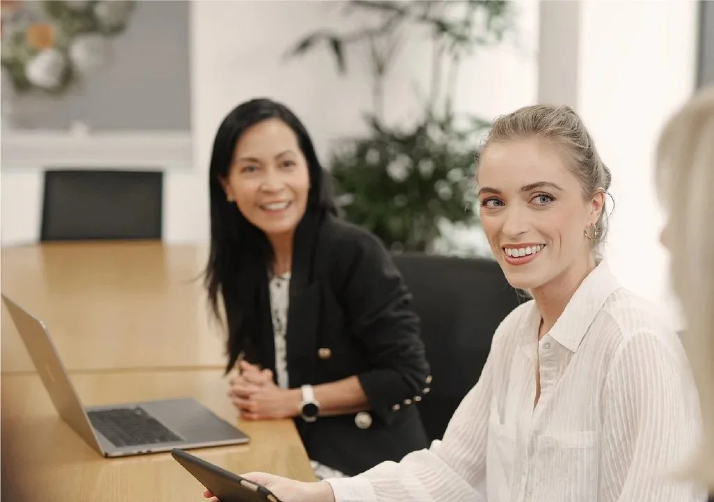 Two women sitting at a conference table, smiling, with one holding a smartphone and the other near a laptop, in a meeting room.