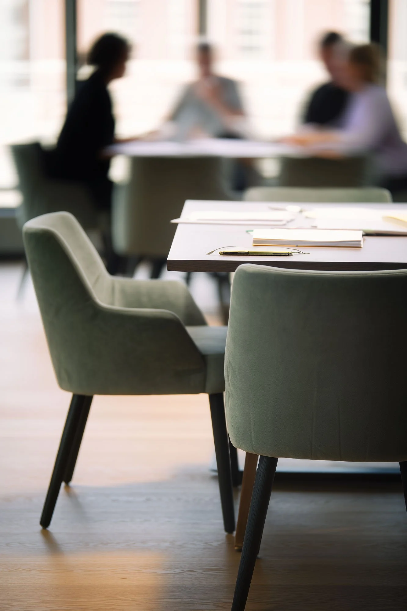 Empty conference room with chairs around a table and documents with a pen on top, with a blurred group of four women in discussion in the background.