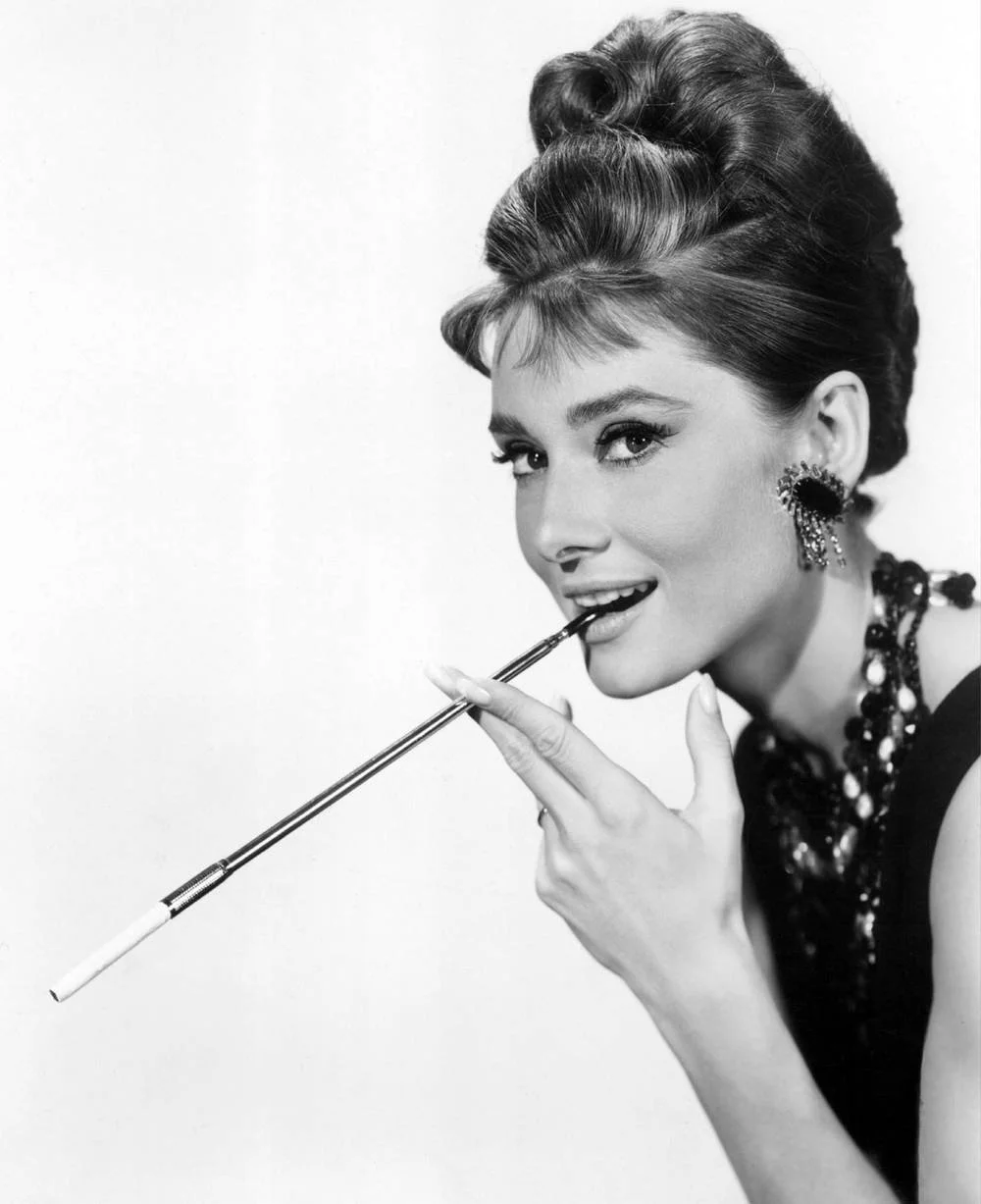 Black and white image of a woman smiling and posed holding a cigarette holder near her mouth, wearing large earrings and a necklace.