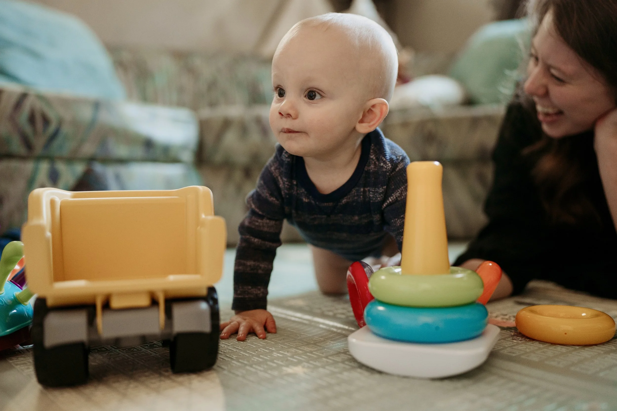 Young baby crawling on a carpeted floor with toys, including a yellow toy dump truck and a colorful stacking ring toy, while a woman smiles nearby.