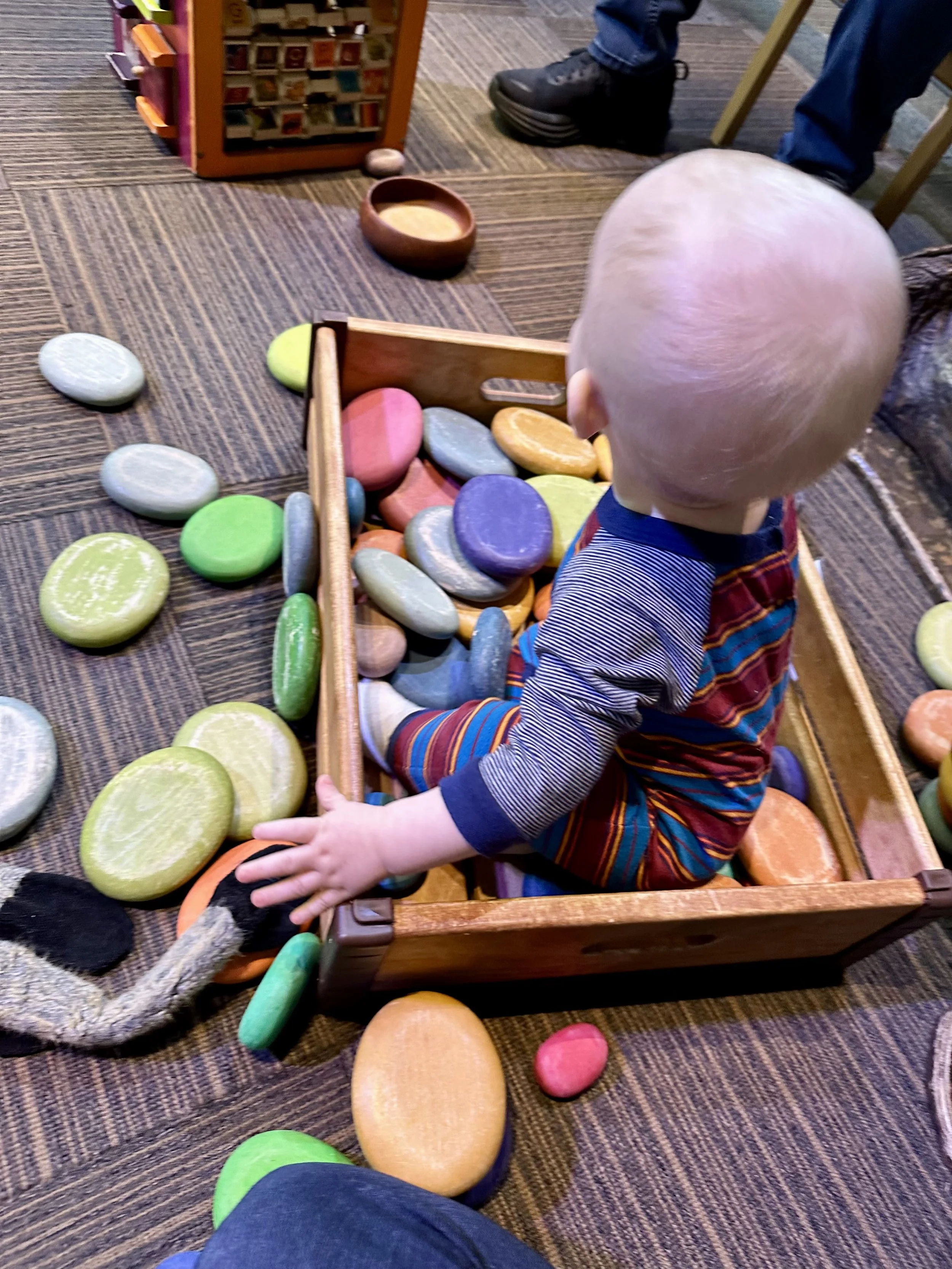 A young child with a bald head sitting in a wooden toy box filled with colorful foam rocks, surrounded by additional foam rocks scattered on the carpeted floor.