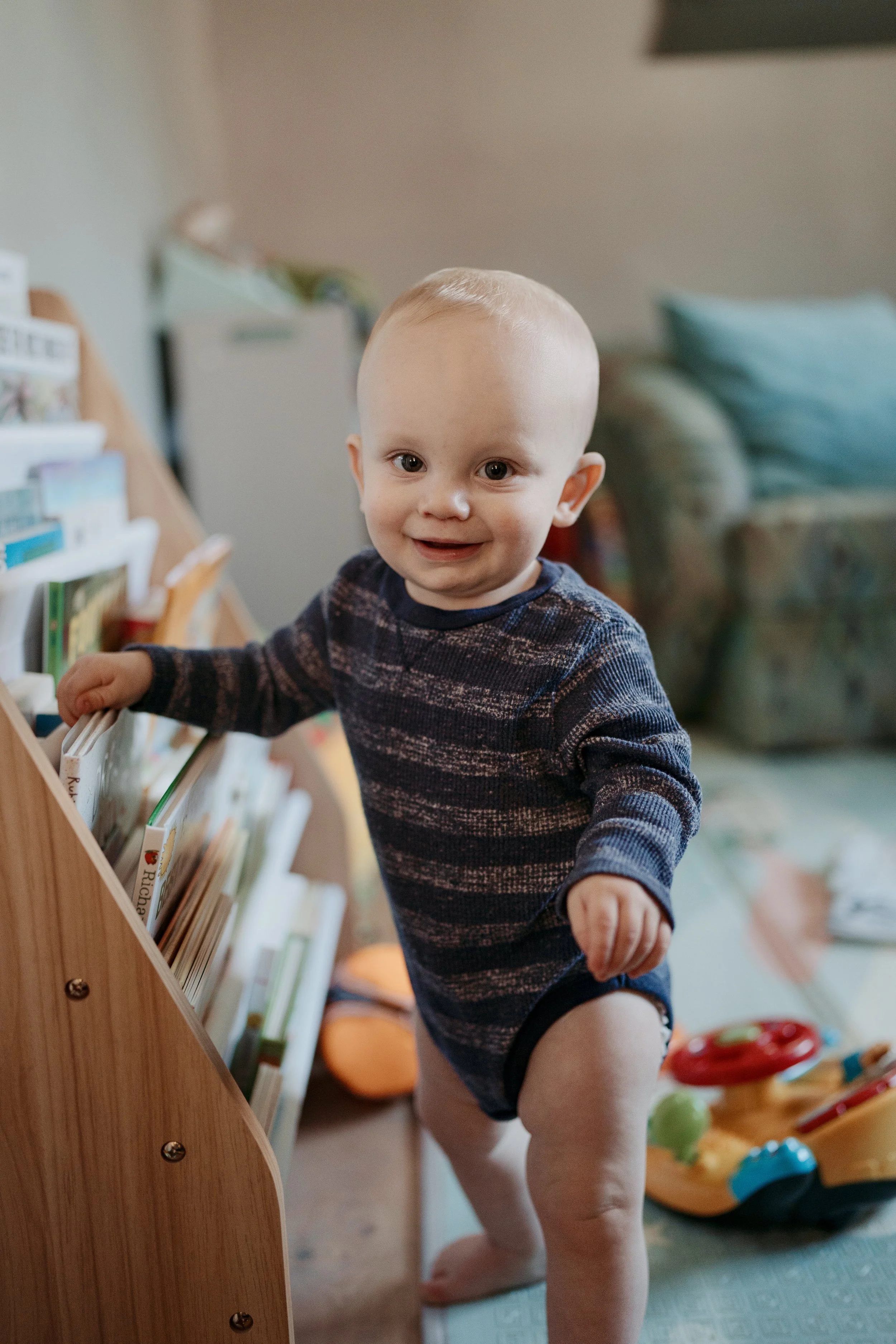 A smiling baby boy with light skin and short hair, in a striped sweater, browsing a small bookshelf indoors.