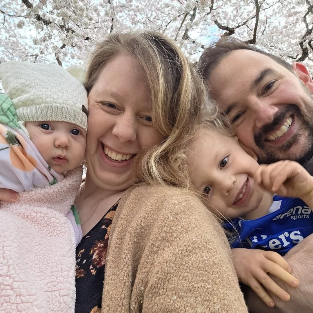 A family of four smiling together outdoors with blooming cherry blossoms in the background.