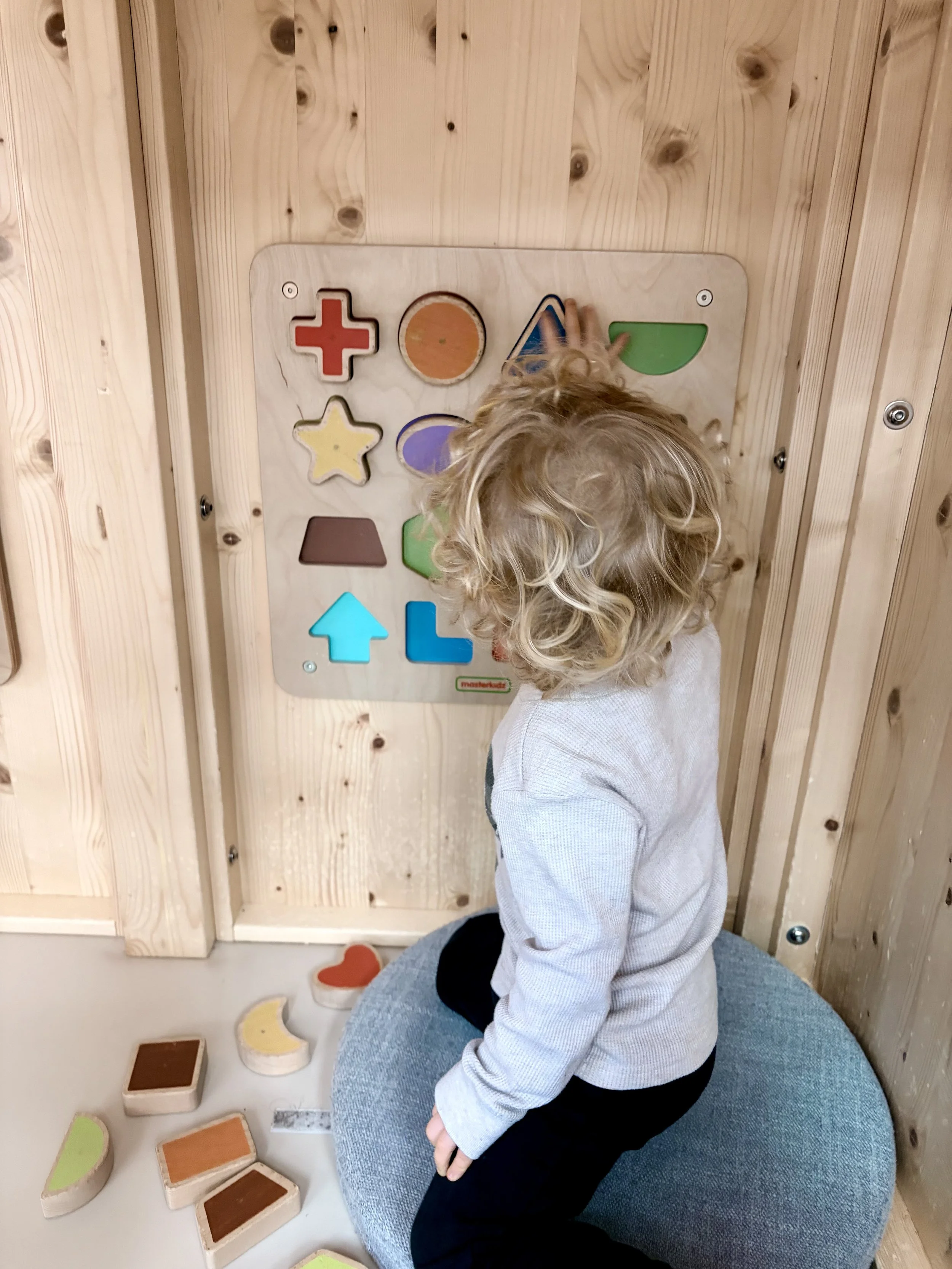 A young child with curly blonde hair interacts with a colorful shape-sorting toy on a wooden wall inside a room. The child is kneeling on a blue cushion.