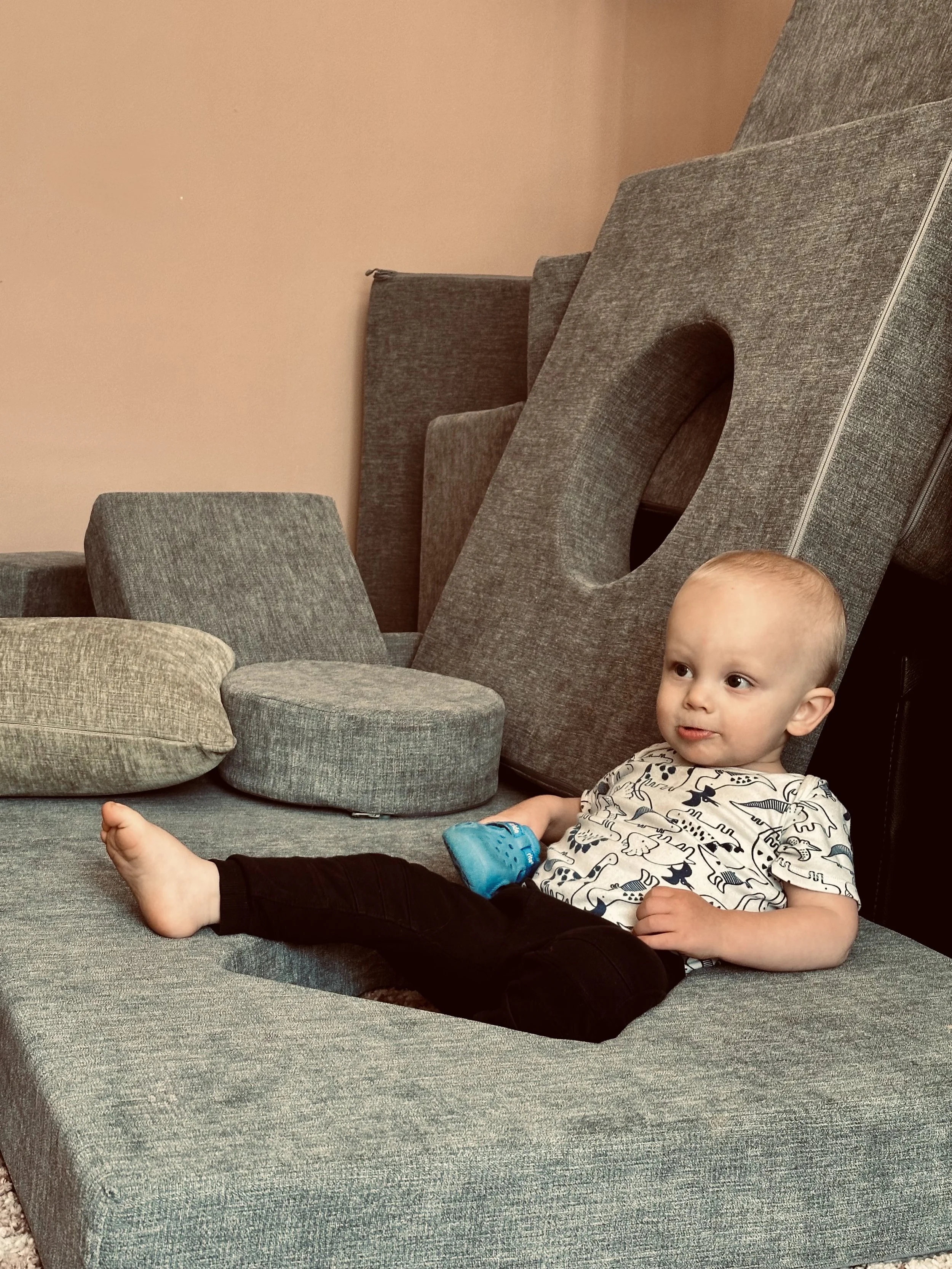 A young child with a shaved head is sitting on a gray sofa, holding a blue toy, with gray and beige furniture pieces and a pinkish wall in the background.