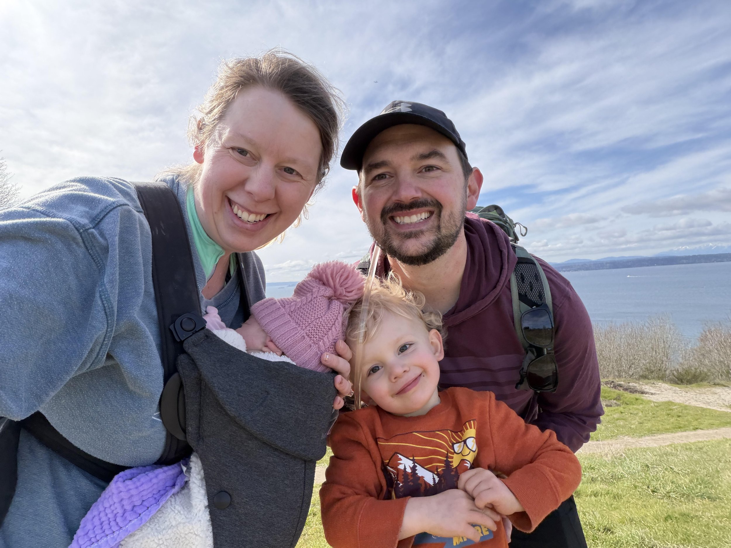 A family of four outdoors near a body of water, smiling at the camera. The mother holds a sleeping baby in a pink hat, while the father and a young boy with a graphic sweatshirt stand beside them. The sky is partly cloudy.