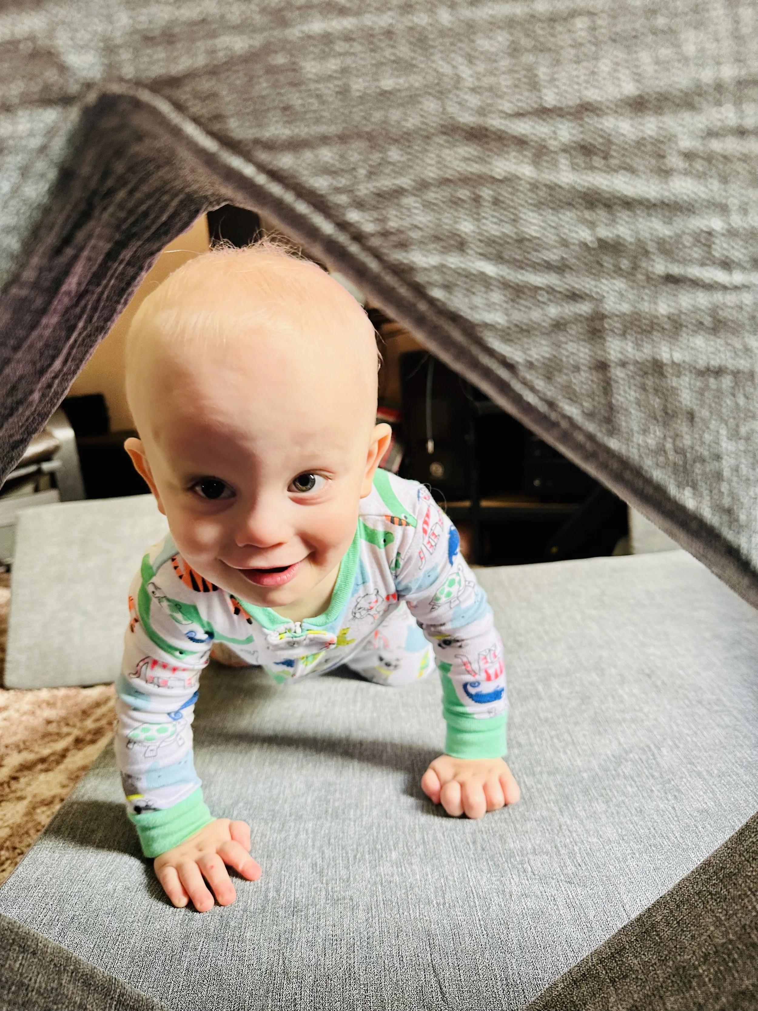 Young child crawling on a gray cushioned surface, viewed through a furniture gap, smiling at the camera.