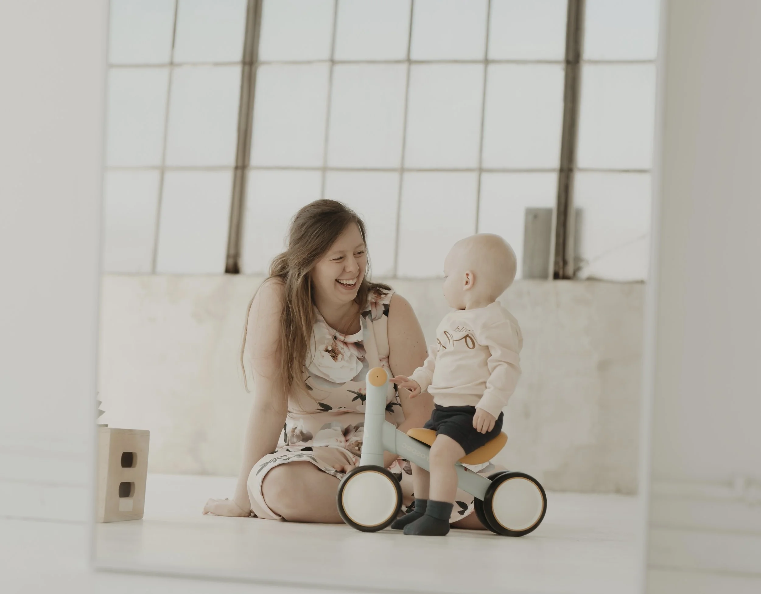 A woman and a young child smiling and playing with a small bicycle in an indoor setting with large windows.
