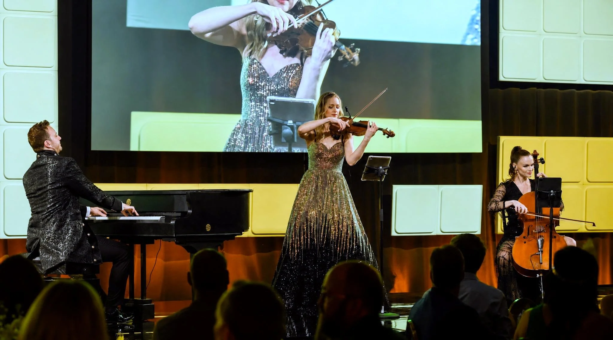 A musical performance with a pianist, two violinists, and a cellist on stage. The main female violinist in a glittery dress is prominently featured, with a large screen behind her showing a close-up of her playing.  Pianist Reuel and the Reuel Trio