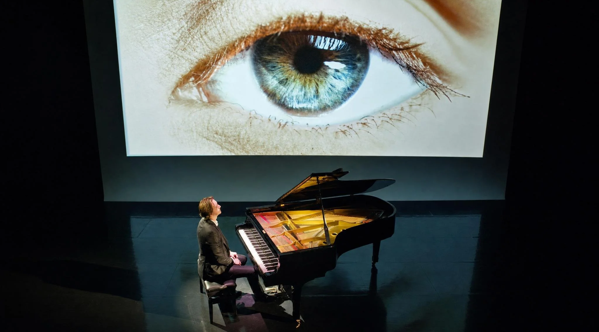 Reuel playing a grand piano on stage with a large screen behind showing a close-up image of a human eye.