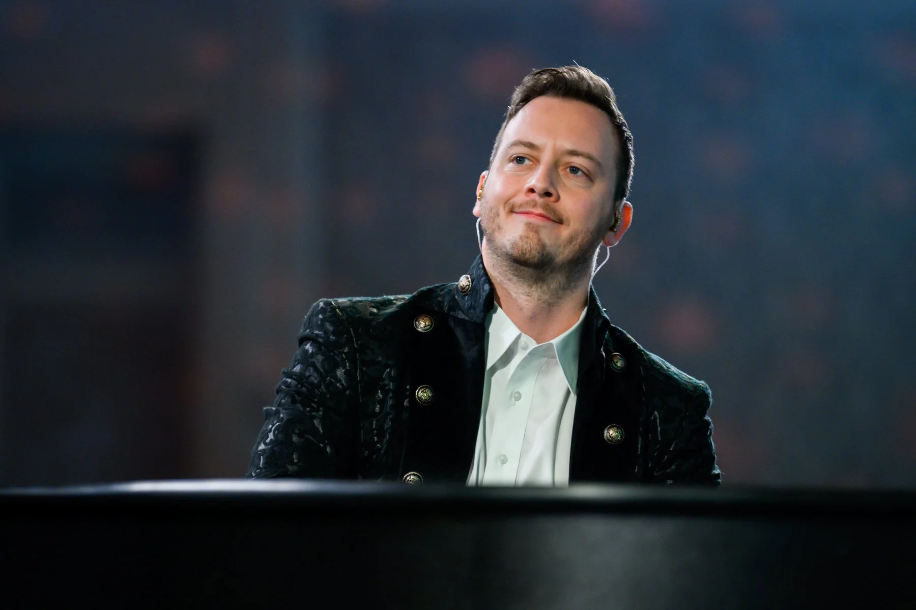 A young man with short dark hair, wearing a white shirt and black jacket with gold buttons, smiling and looking slightly to the right. The background is blurred.