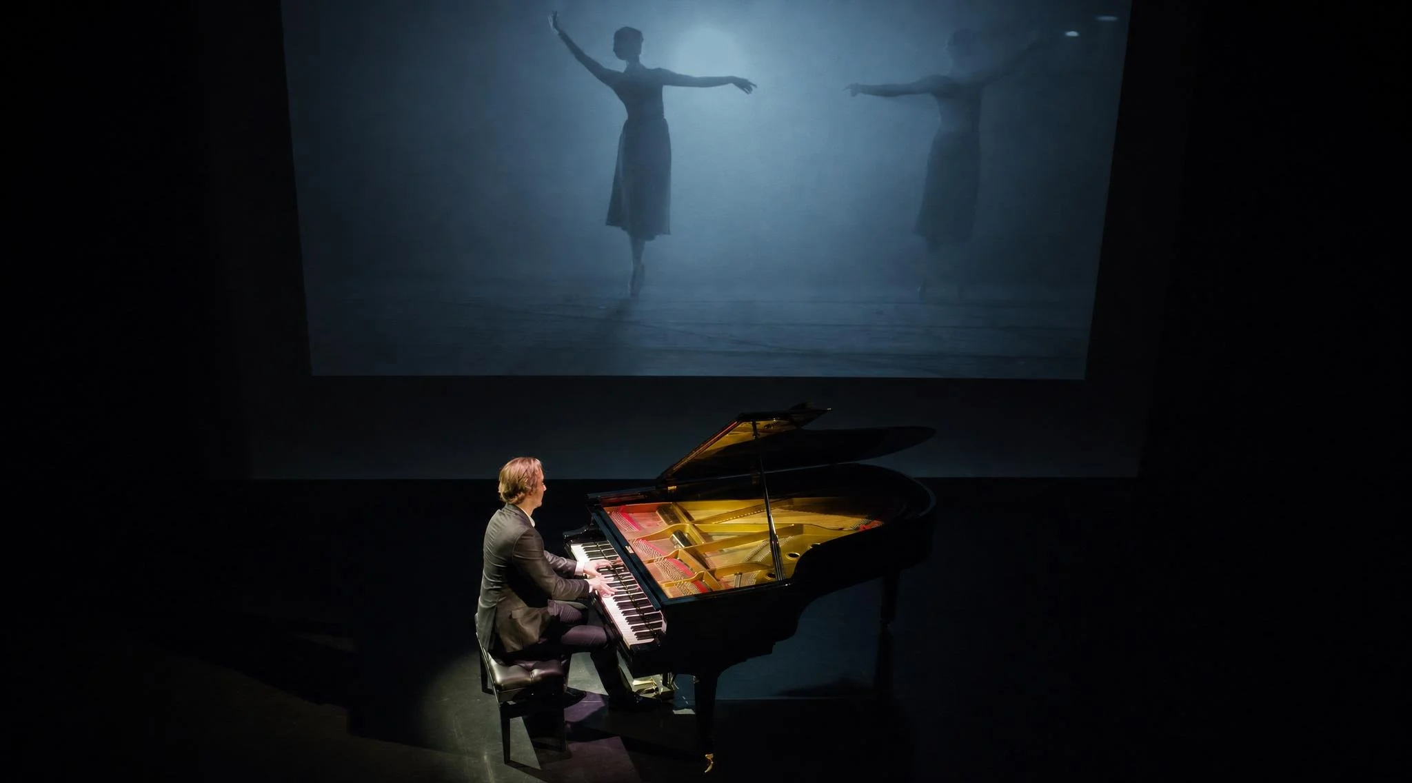 Reuel playing a grand piano on stage with a large screen behind him displaying a ballet dancer in an elegant pose.