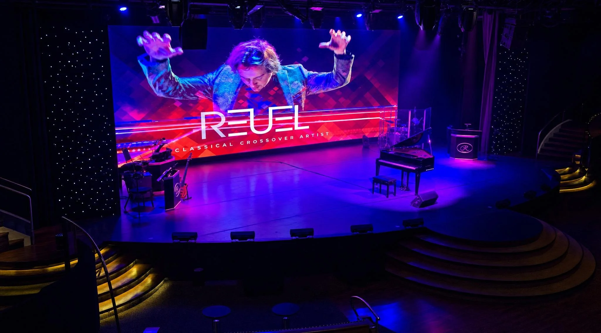 Empty stage with a large LED screen displaying an artist named Reuel with the subtitle 'Classical Crossover Artist.' On the stage are musical instruments, including a grand piano and guitars, with purple and blue stage lighting.