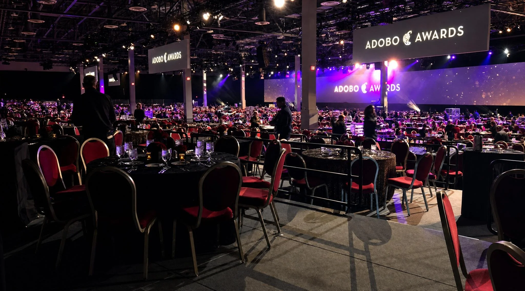 Large conference hall set up for an awards event with round tables covered in black tablecloths and red chairs. Multiple large screens above display the 'ADOBE AWARDS' logo. The stage at the front is lit with purple and yellow lighting.  Pianist Reue