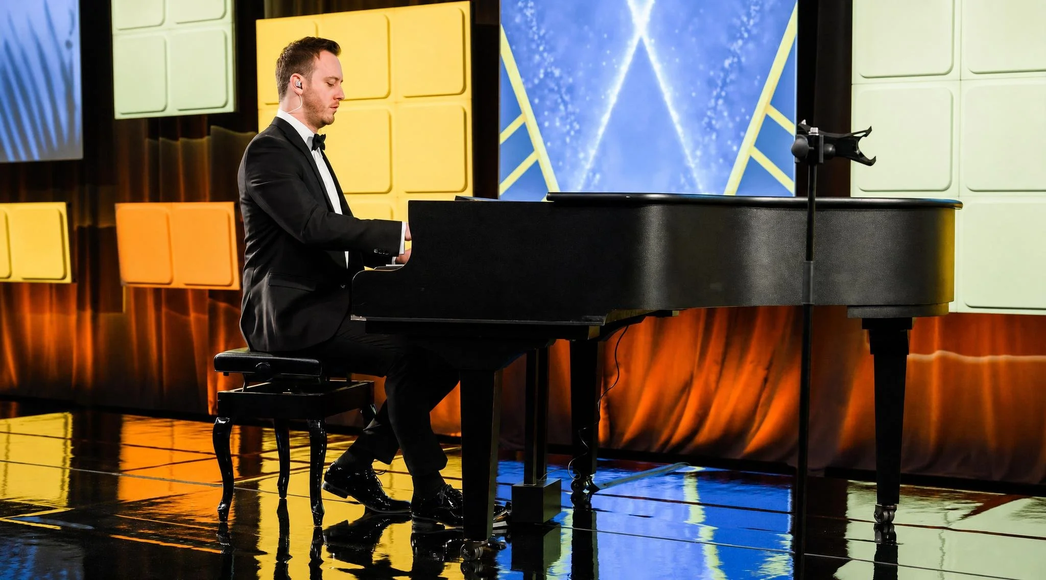 Reuel in a black tuxedo playing a grand piano on a brightly lit stage with colorful backdrop panels.