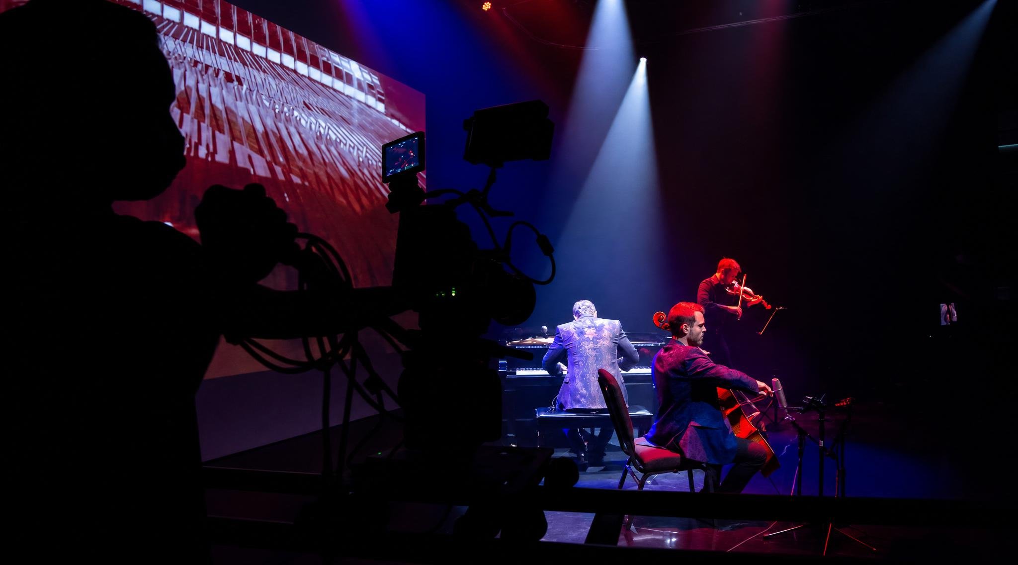 Reuel Trio perform on stage with a pianist, violinist, and cellist, illuminated by stage lights, with a large screen displaying abstract visuals in the background.