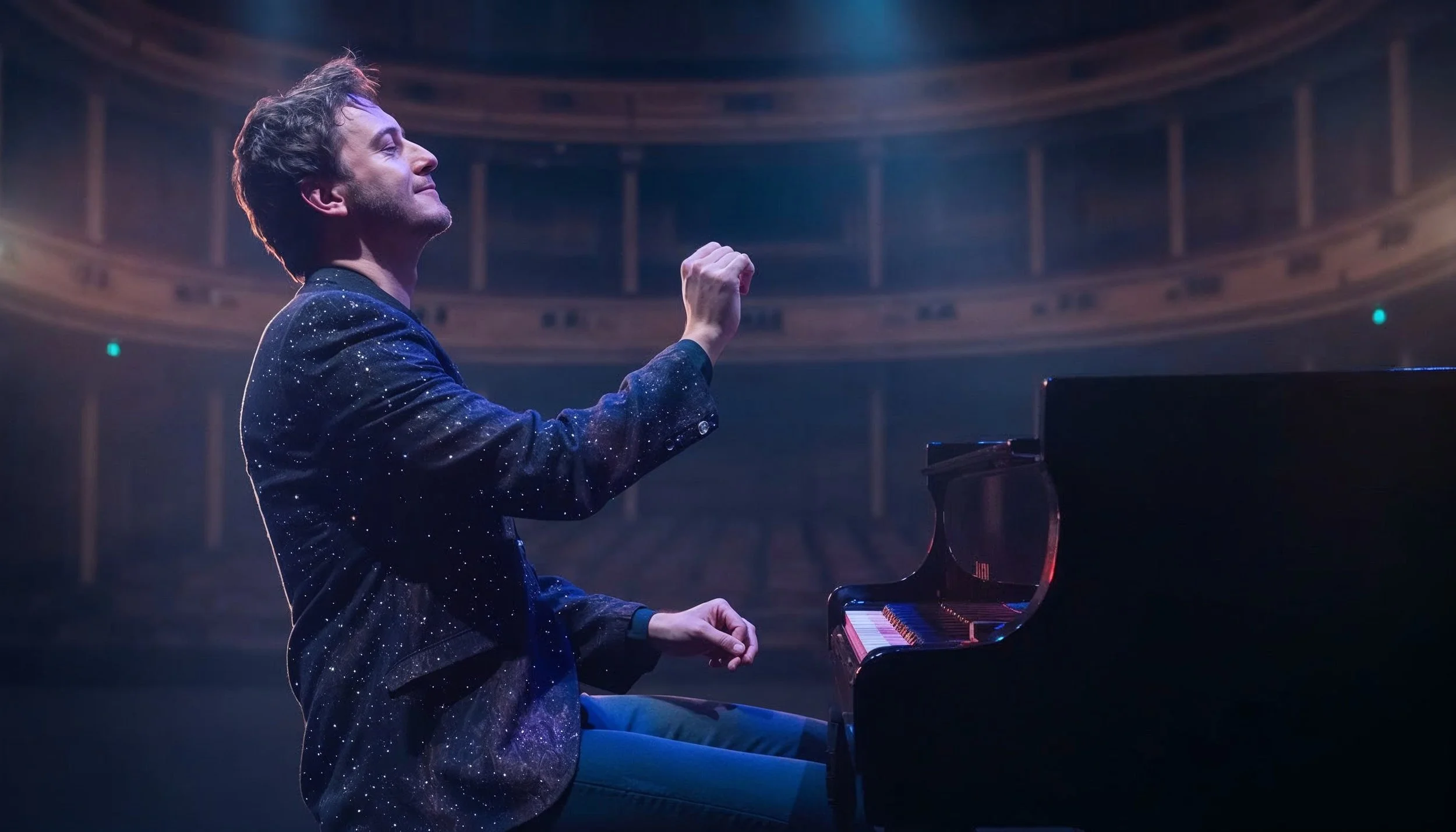 A man wearing a starry-patterned suit playing the piano on stage with a spotlight shining on him in an auditorium.