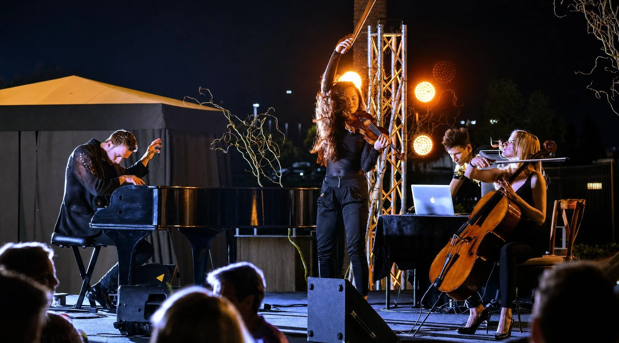 Musicians performing outdoors at night, including a pianist, a violinist, and a cellist, with a woman conducting or leading.   Pianist Reuel