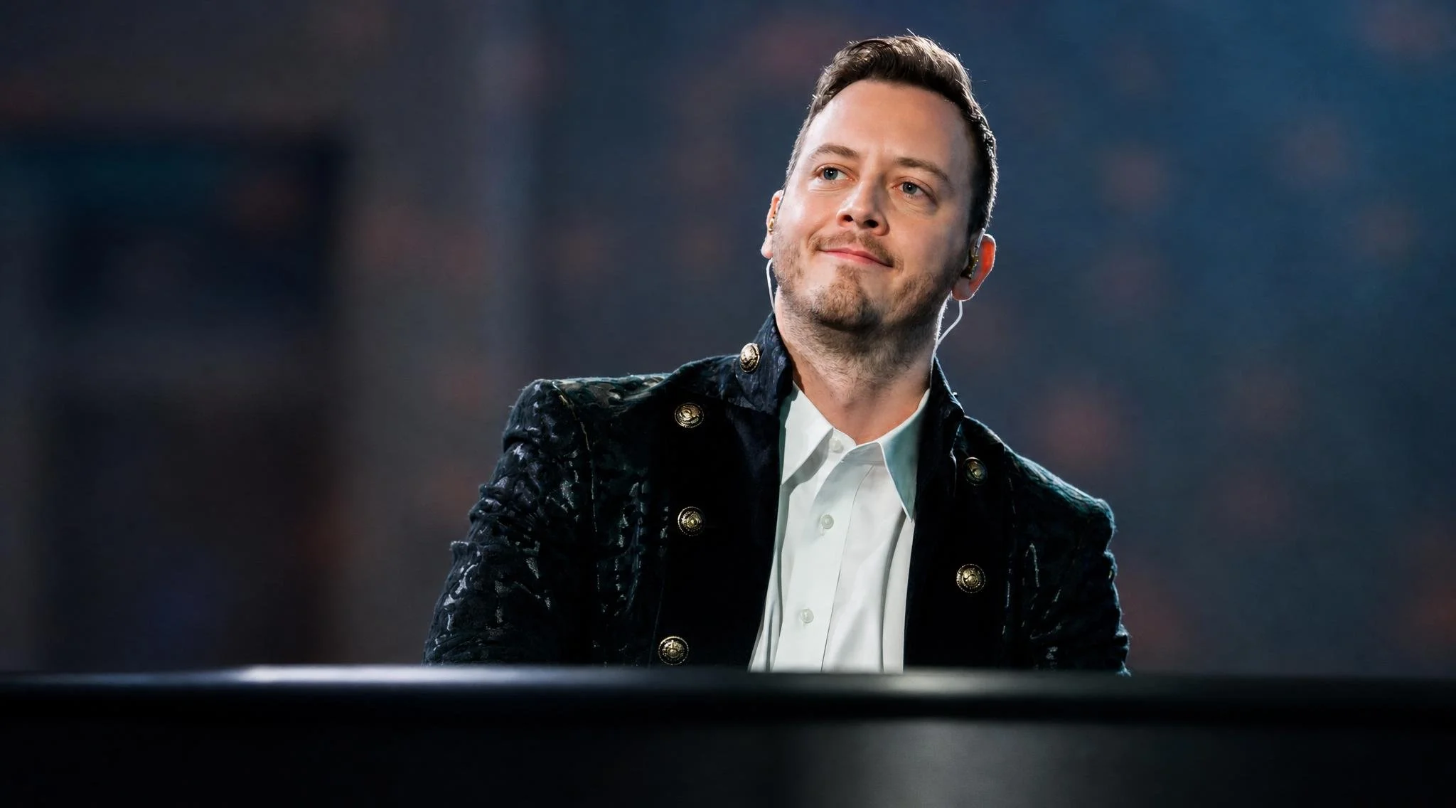 Reuel with light brown hair and a short beard, wearing a white shirt and a dark velvet jacket with gold buttons, is smiling and looking slightly to the side.