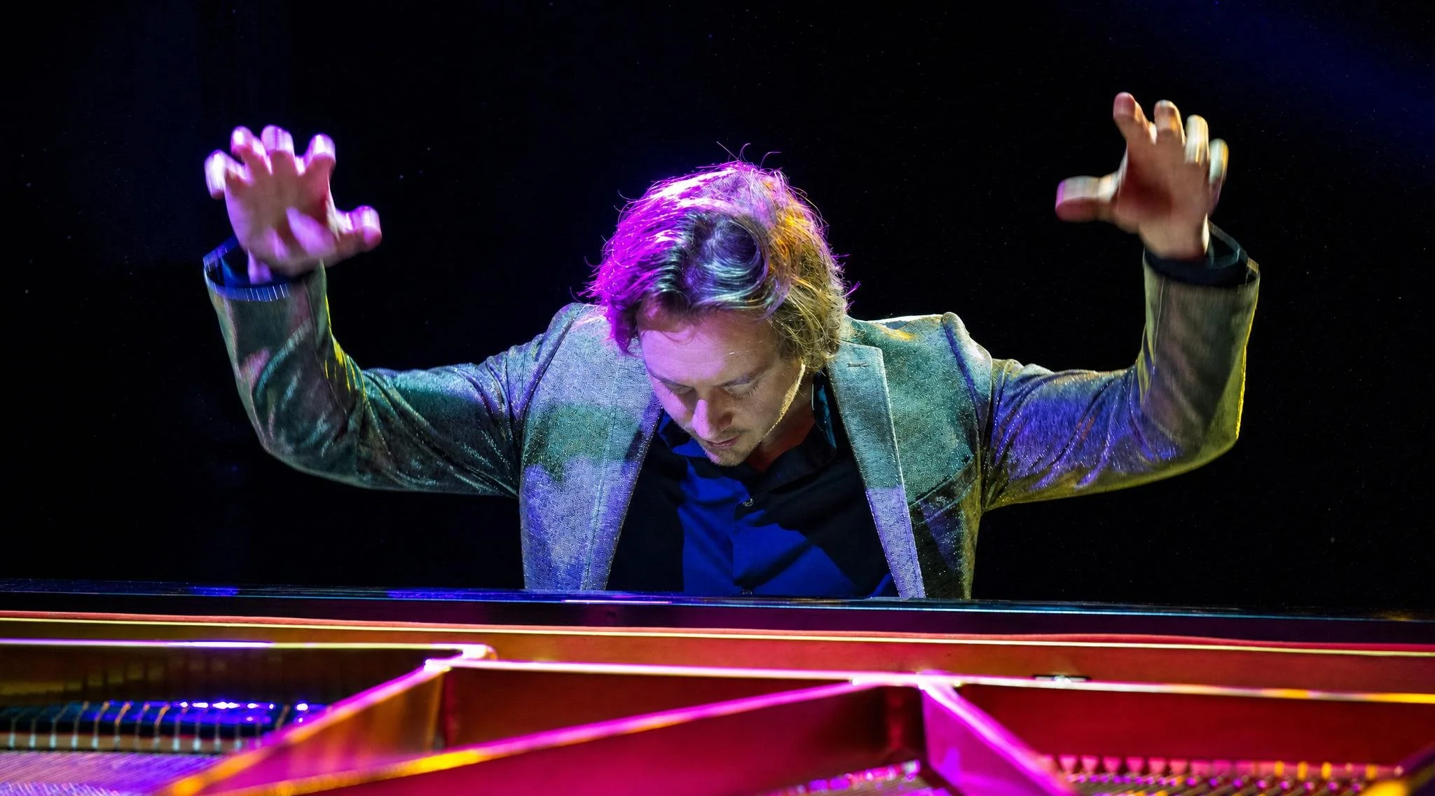 Reuel playing a grand piano with his head bowed and hands raised in the air against a dark background.