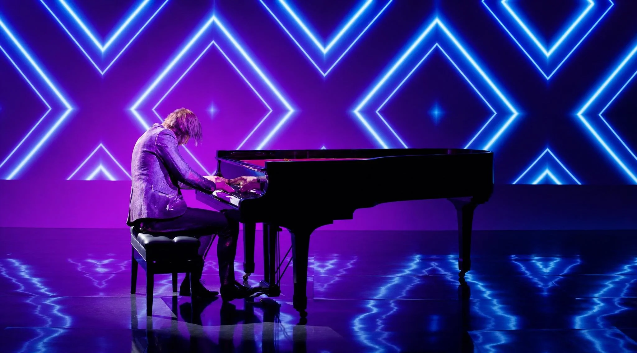 Reuel playing a grand piano on a stage with a neon-lit geometric background.