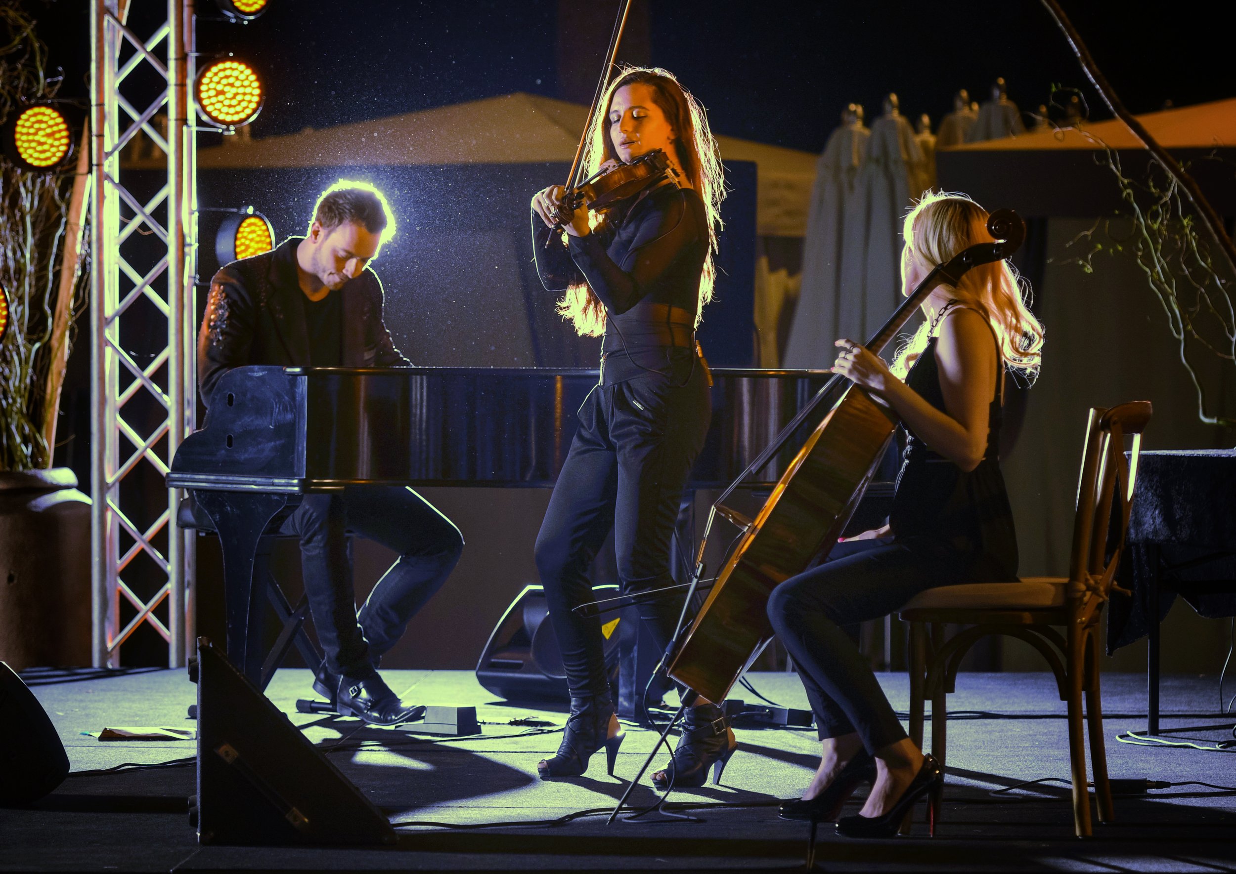 Three musicians performing on stage at night, one playing the piano and two playing violins, illuminated by stage lights.