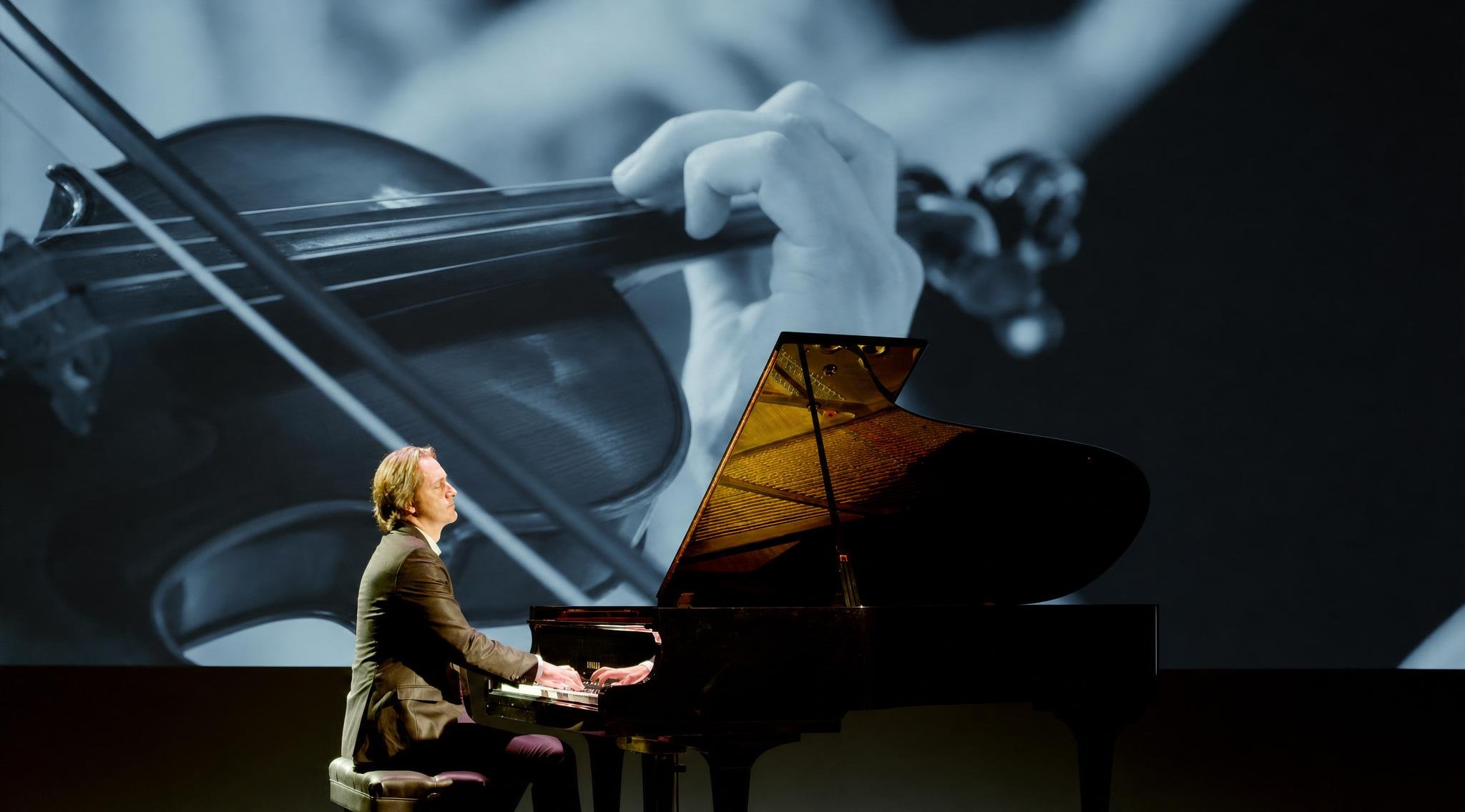 Reuel playing a grand piano on a stage with a large projected image of a musician playing a violin in the background.