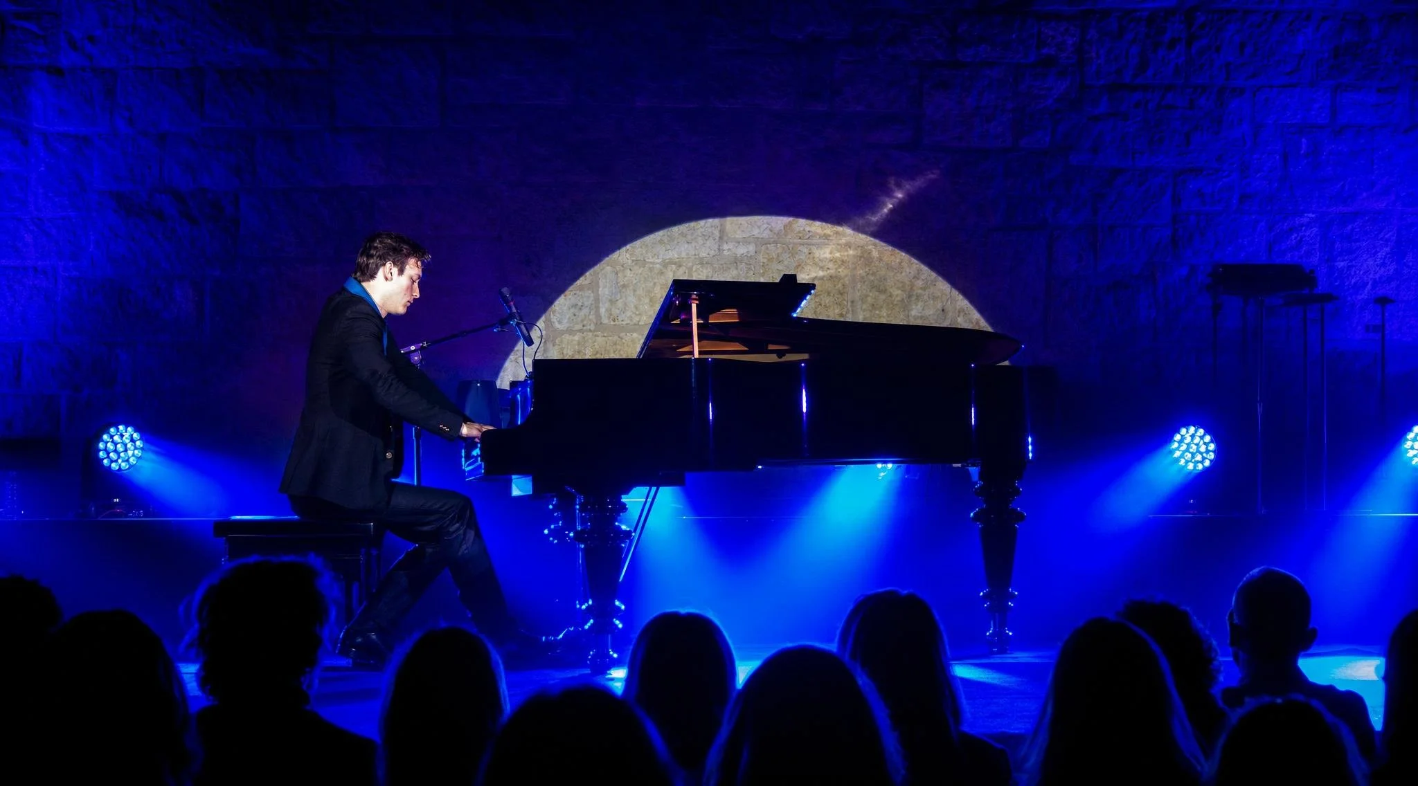 Reuel in a black jacket playing a grand piano on stage with blue lighting, in front of an audience.