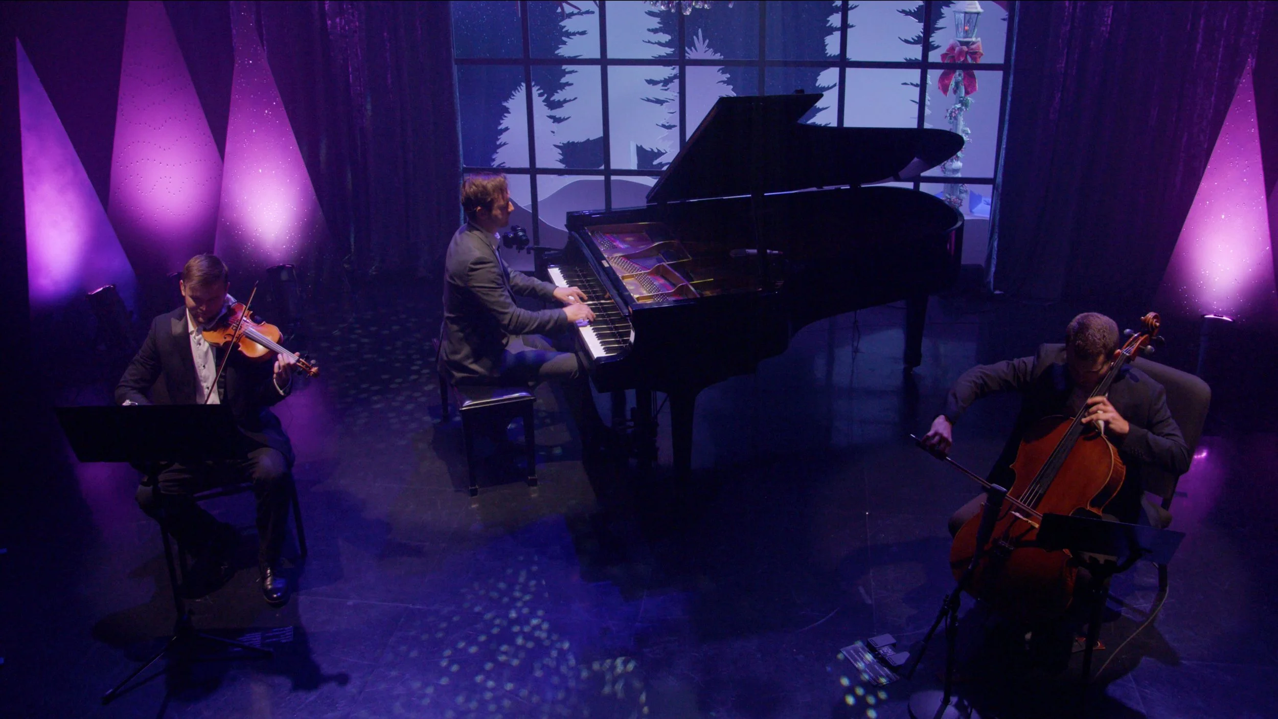 Reuel Trio performing on a stage with purple lighting, featuring a pianist, a violinist, and a cellist, with a winter-themed window backdrop.