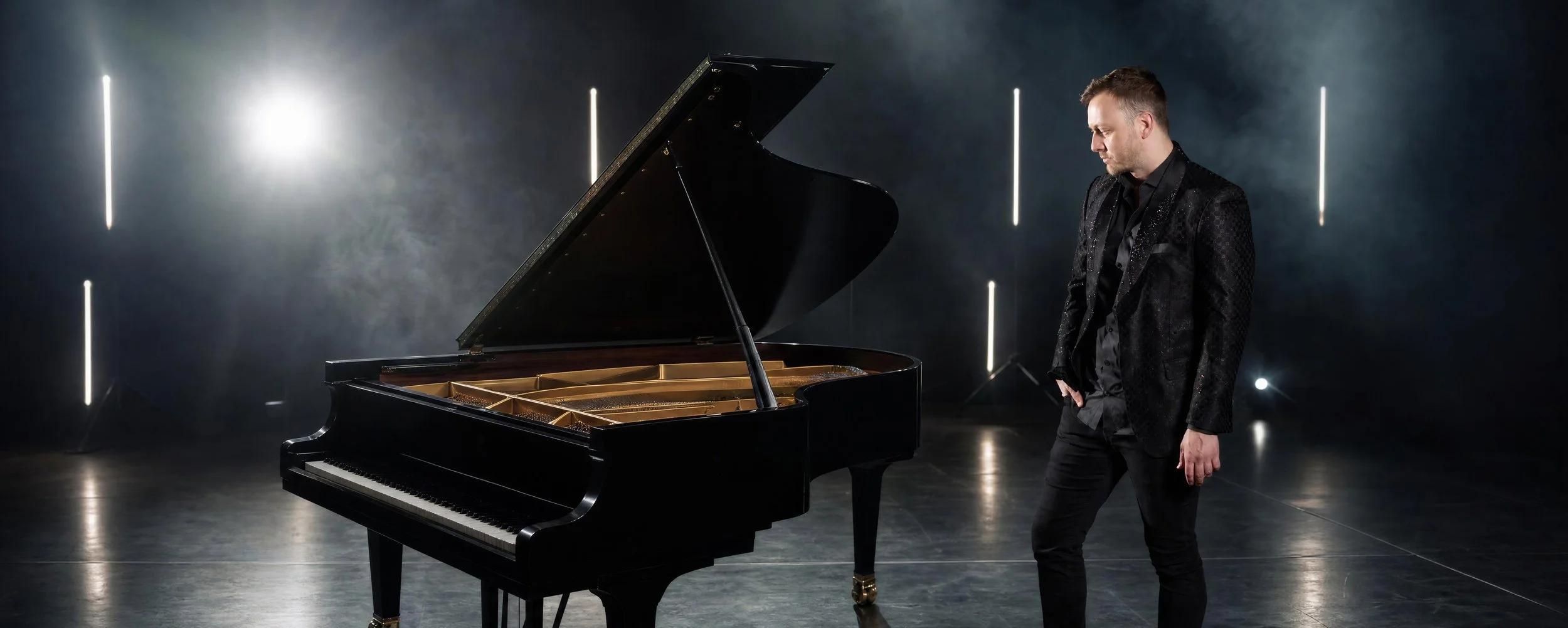 A man in a black suit stands next to a grand piano in a dark, illuminated room.