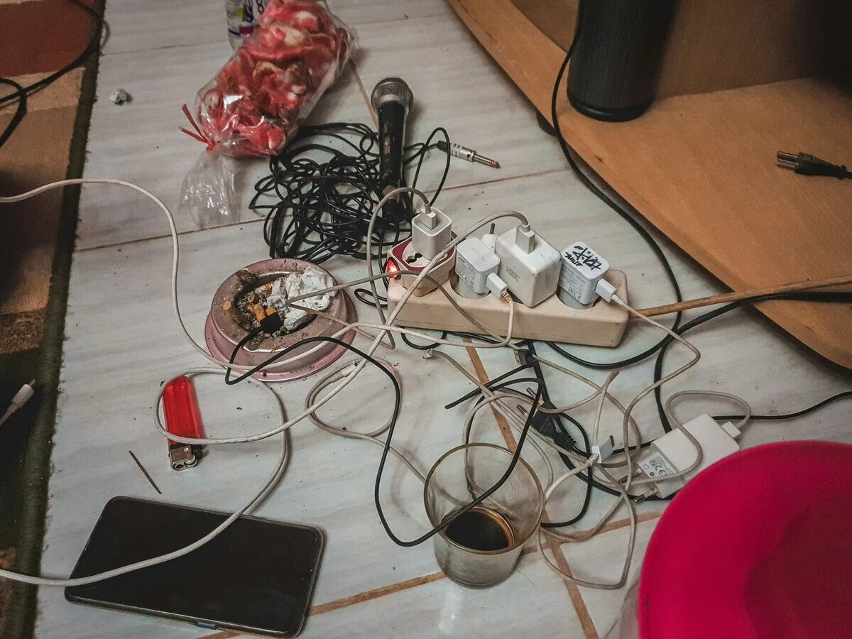 Cluttered floor with tangled electrical cords, a smartphone, a glass of dark coffee, a partially melted smoking ashtray, a micro USB charger, power strips, and a jar of marshmallows.