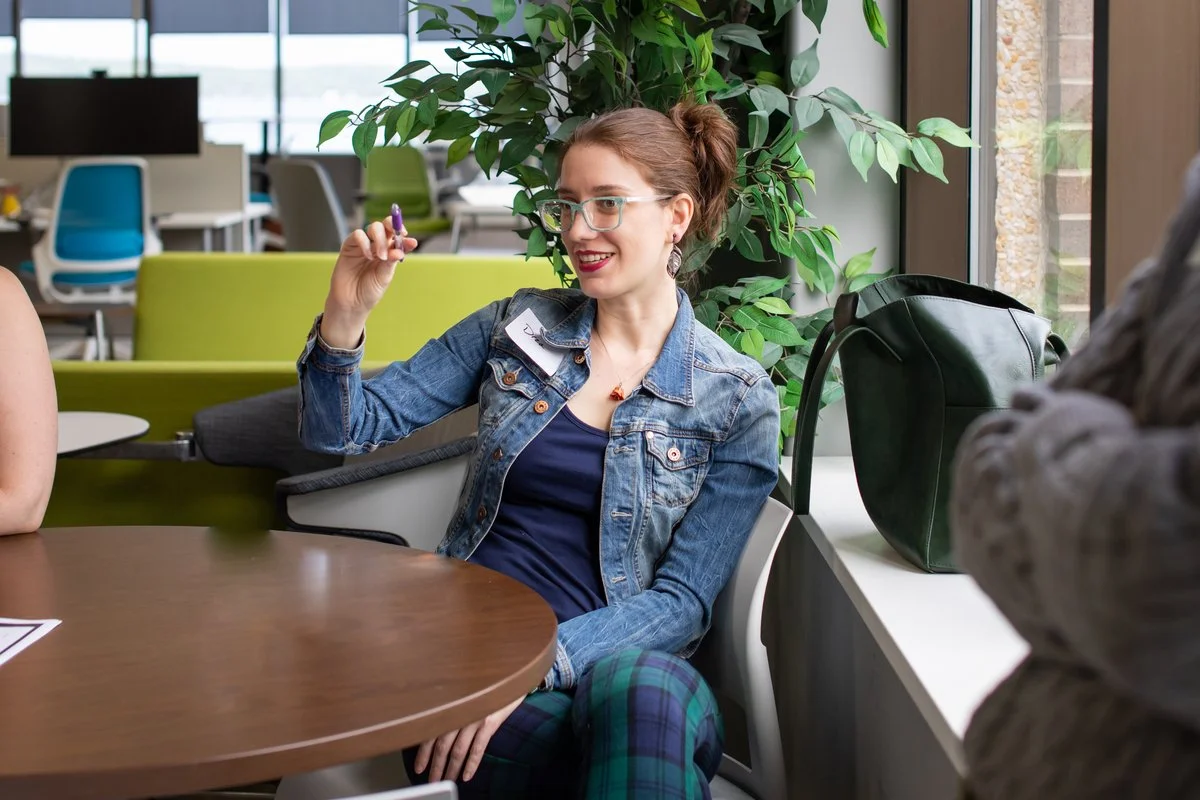 A woman with glasses, red hair in a bun, wearing a denim jacket and plaid pants, sitting at a table in a modern office space, smiling and holding a purple marker, with a large green plant behind her.