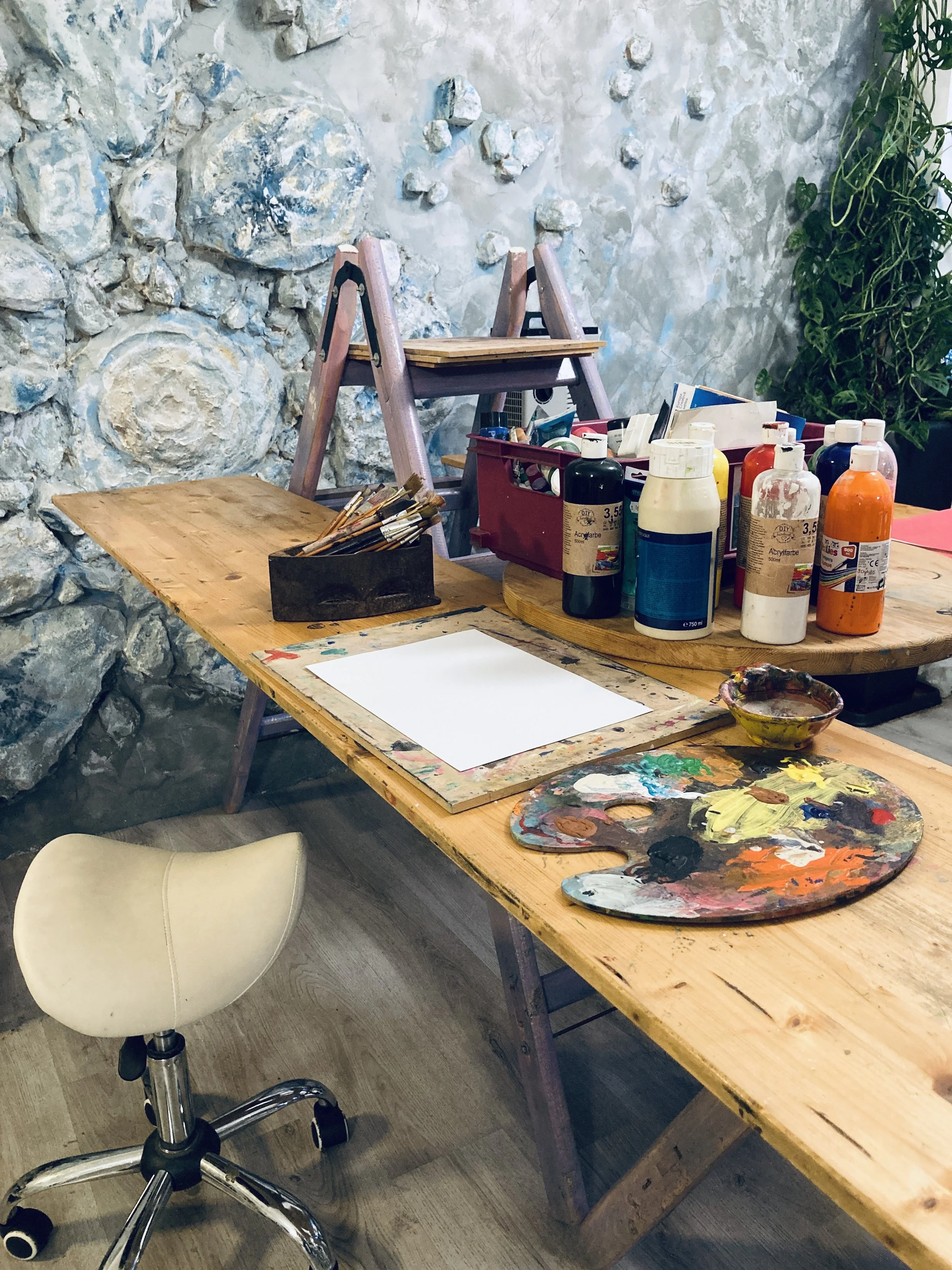 An art studio workspace with a wooden table covered in paint bottles, brushes, a palette, a blank paper, and an easel, set against a painted stone wall and a green plant.